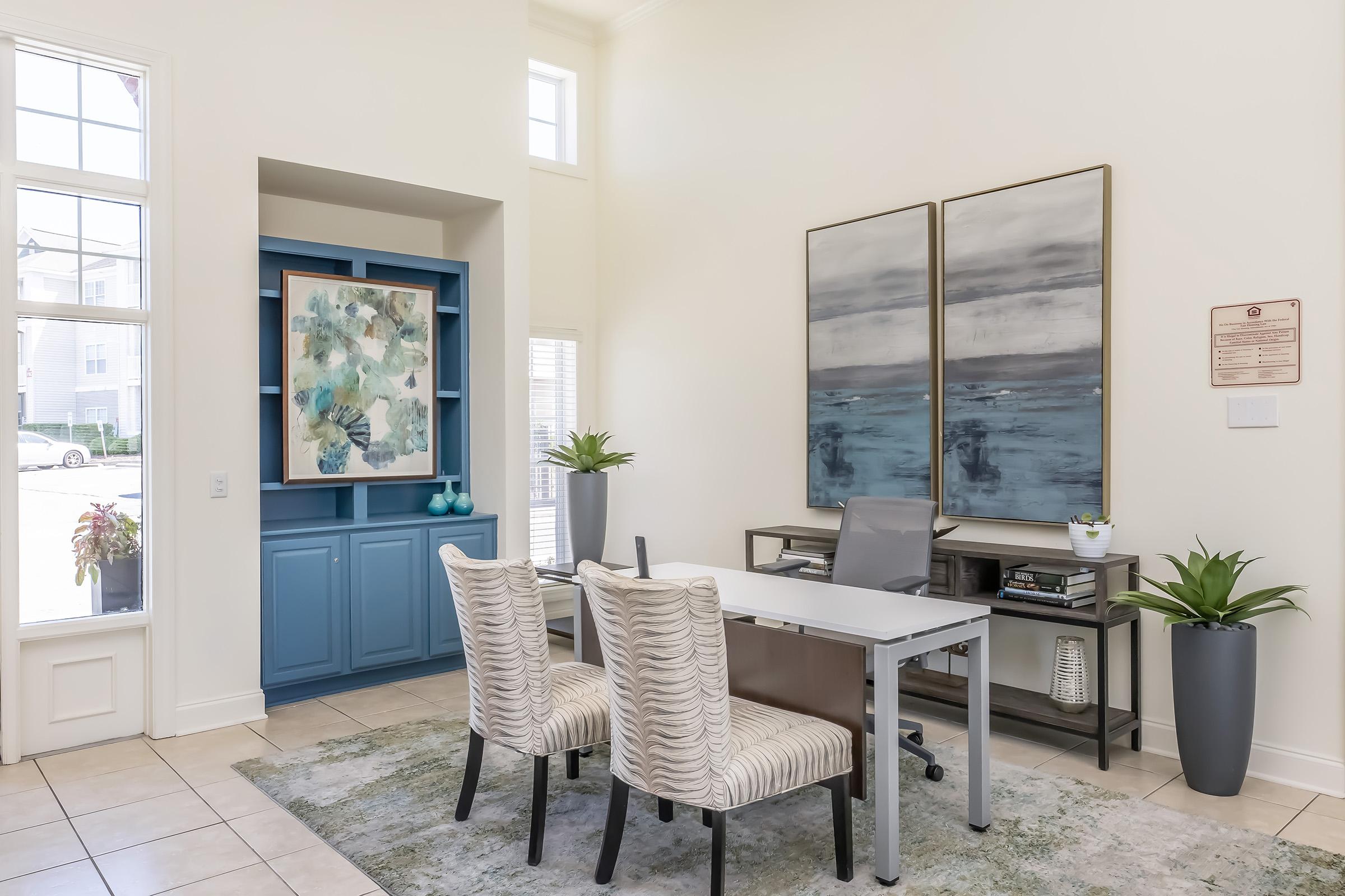 A bright and modern office space featuring a light-colored wall with large windows. There is a gray desk with a chair, flanked by two upholstered chairs. A blue cabinet with decorative artwork and a small plant is displayed against the wall, while a green area rug adds warmth to the room.