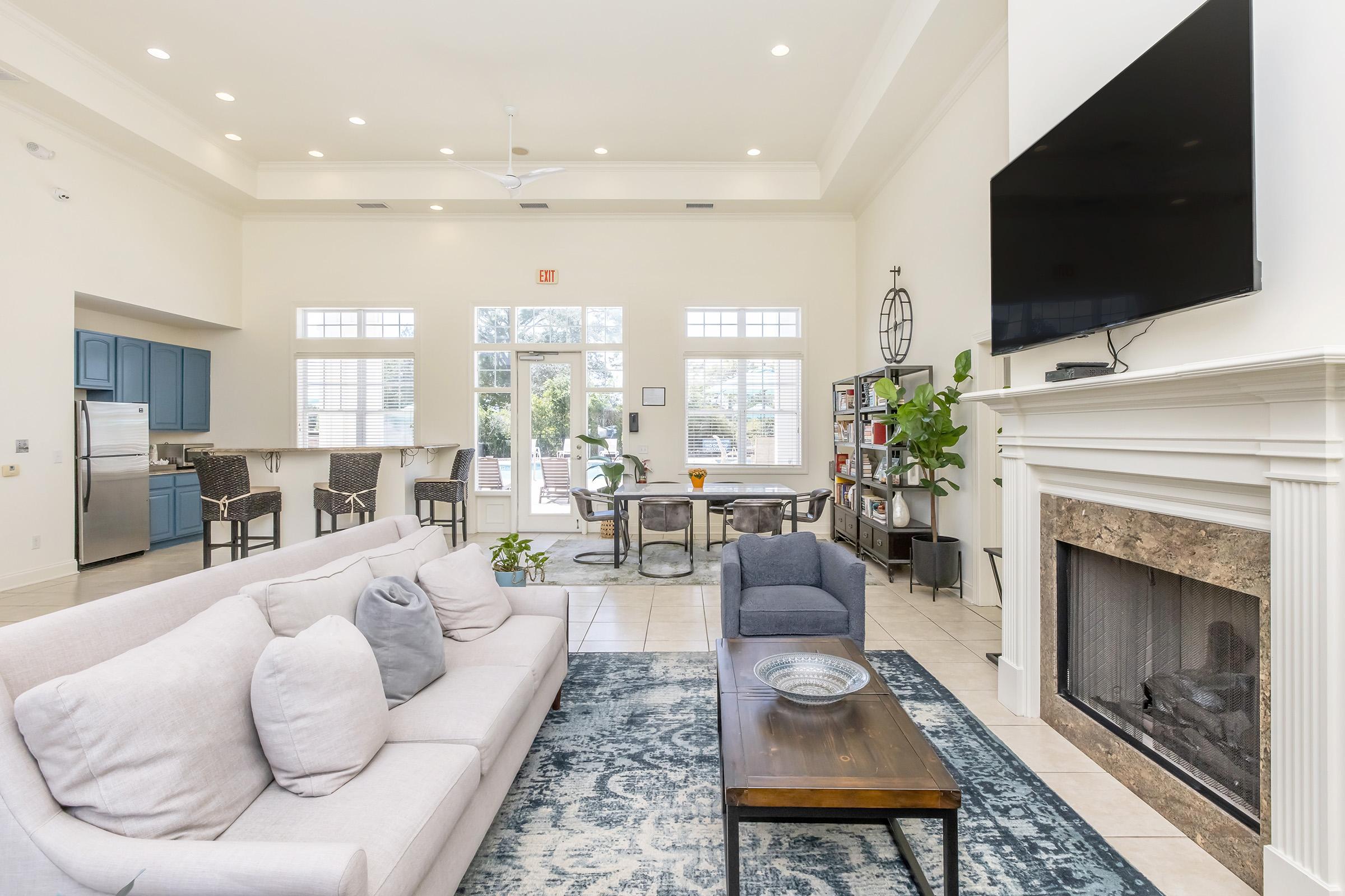 A spacious living room featuring a large beige sofa with decorative pillows, a wooden coffee table, and a cozy reading chair. There is a fireplace with a stone surround and a wall-mounted TV. The room has large windows allowing natural light, and a dining area with a table and chairs in the background.