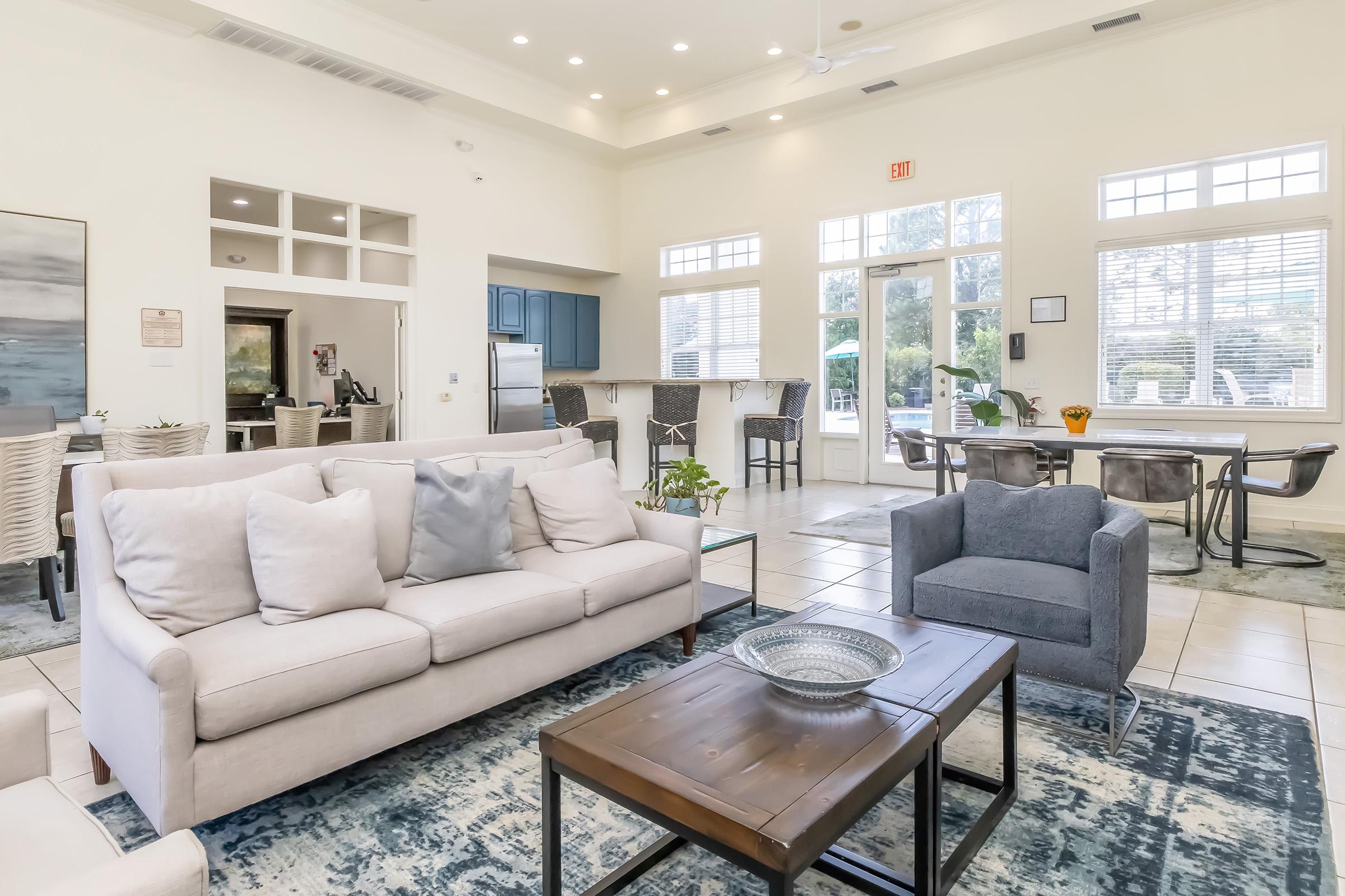 A spacious living area featuring a light-colored couch with decorative pillows, a wooden coffee table, and a cozy armchair. Large windows allow natural light to fill the room. The area includes a dining space with a table set for four, blue cabinets in the kitchen area, and a stylish rug on the floor.