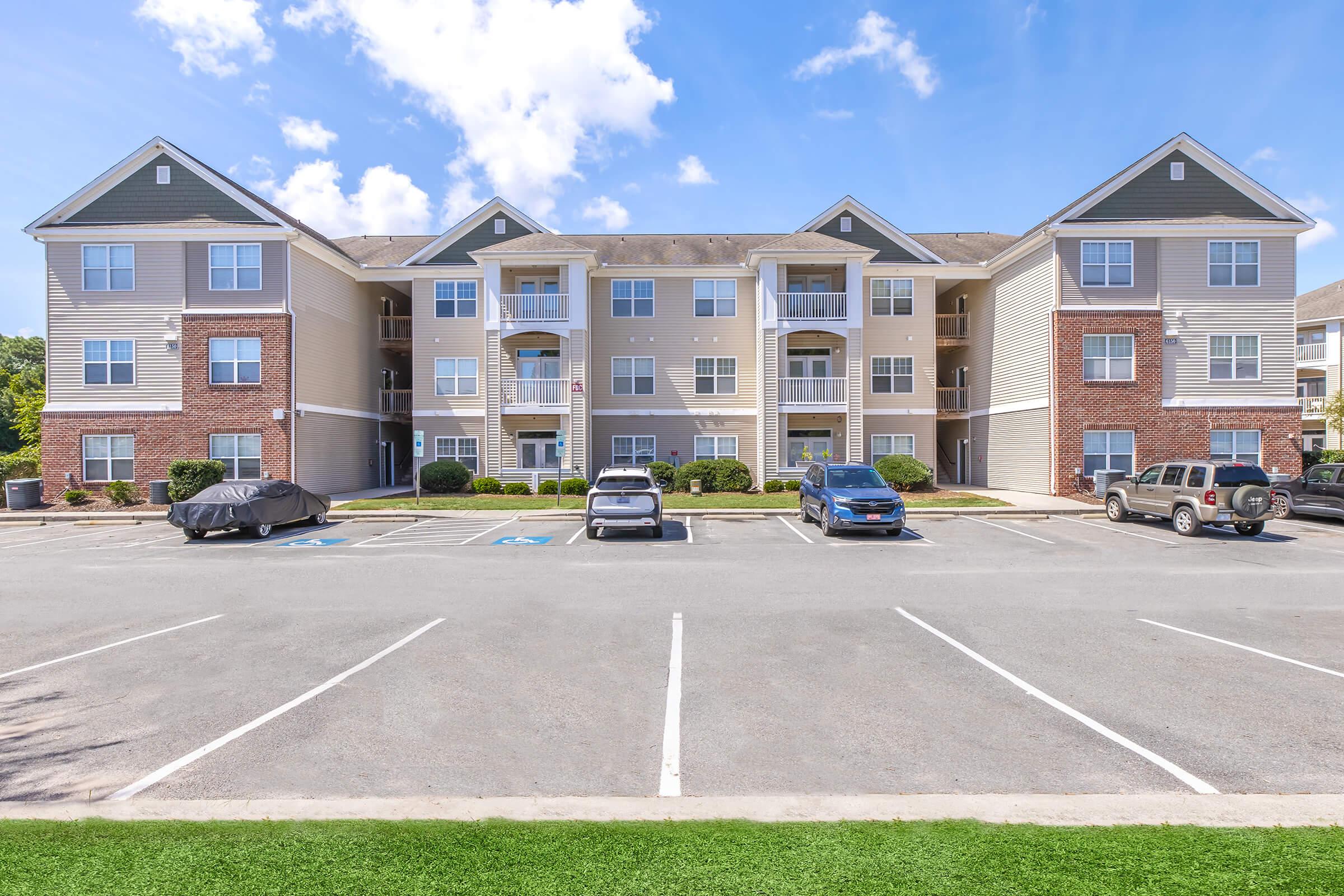 A well-maintained multi-story apartment building with a brick and siding exterior. The building features multiple balconies and windows. In the foreground, there are several parked cars and marked parking spaces, with green landscaping visible. The sky is clear with a few clouds.
