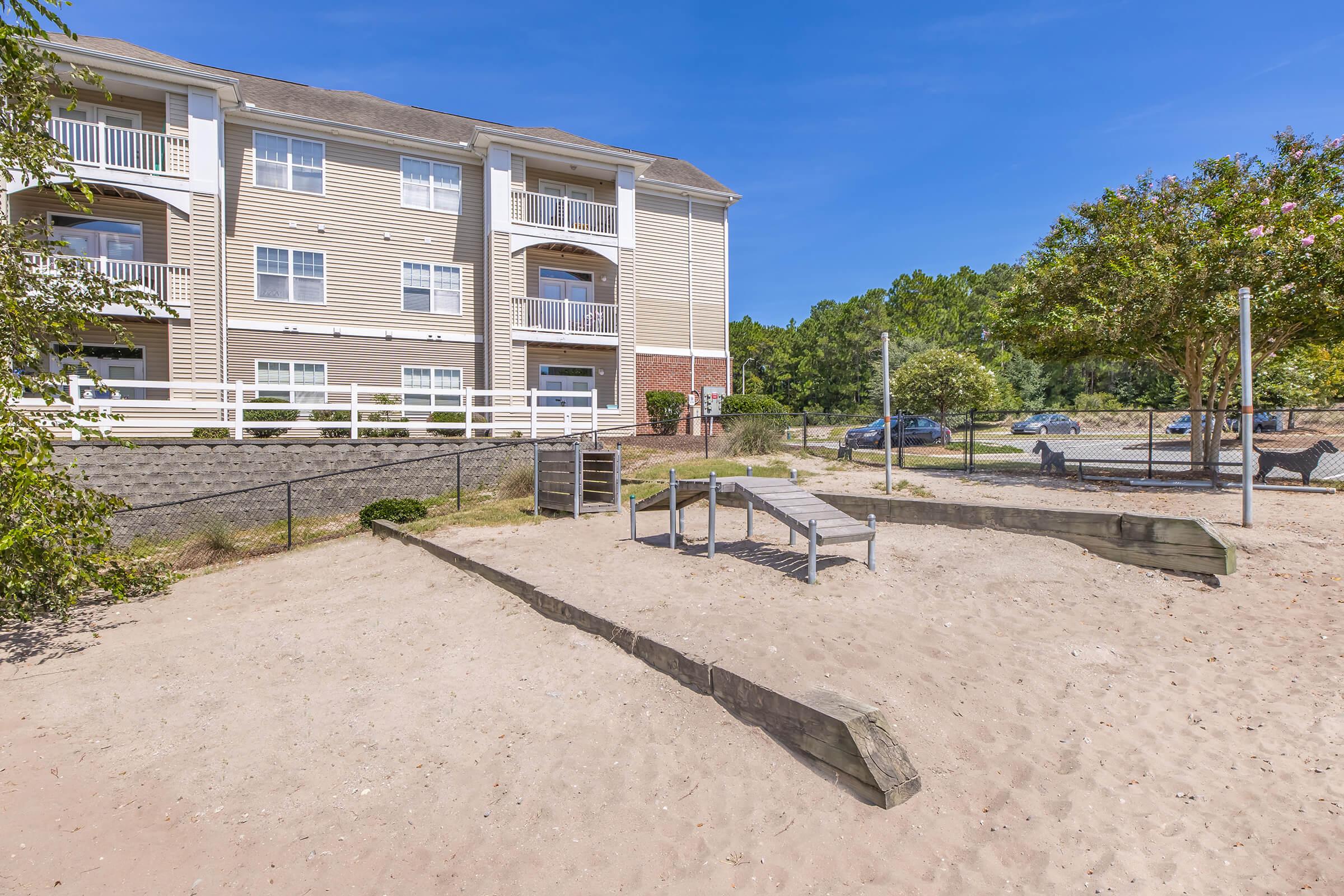 A sandy playground area with equipment, surrounded by a fence. In the background, a three-story residential building is visible, along with trees and blue sky above. Several parked cars are seen nearby, suggesting a community setting.