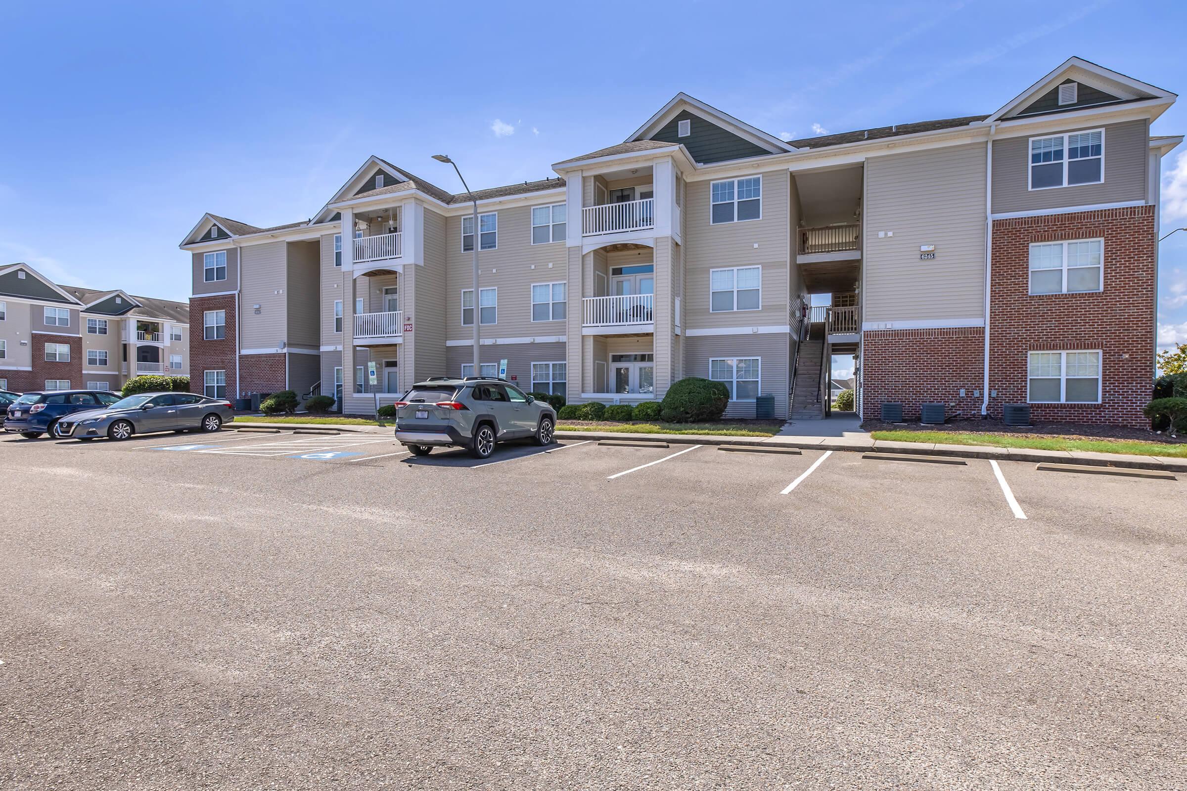 A multi-story apartment building featuring several balconies, surrounded by a parking lot with multiple cars parked. The building exterior has a light beige color with brick accents, landscaping, and clear blue skies in the background.