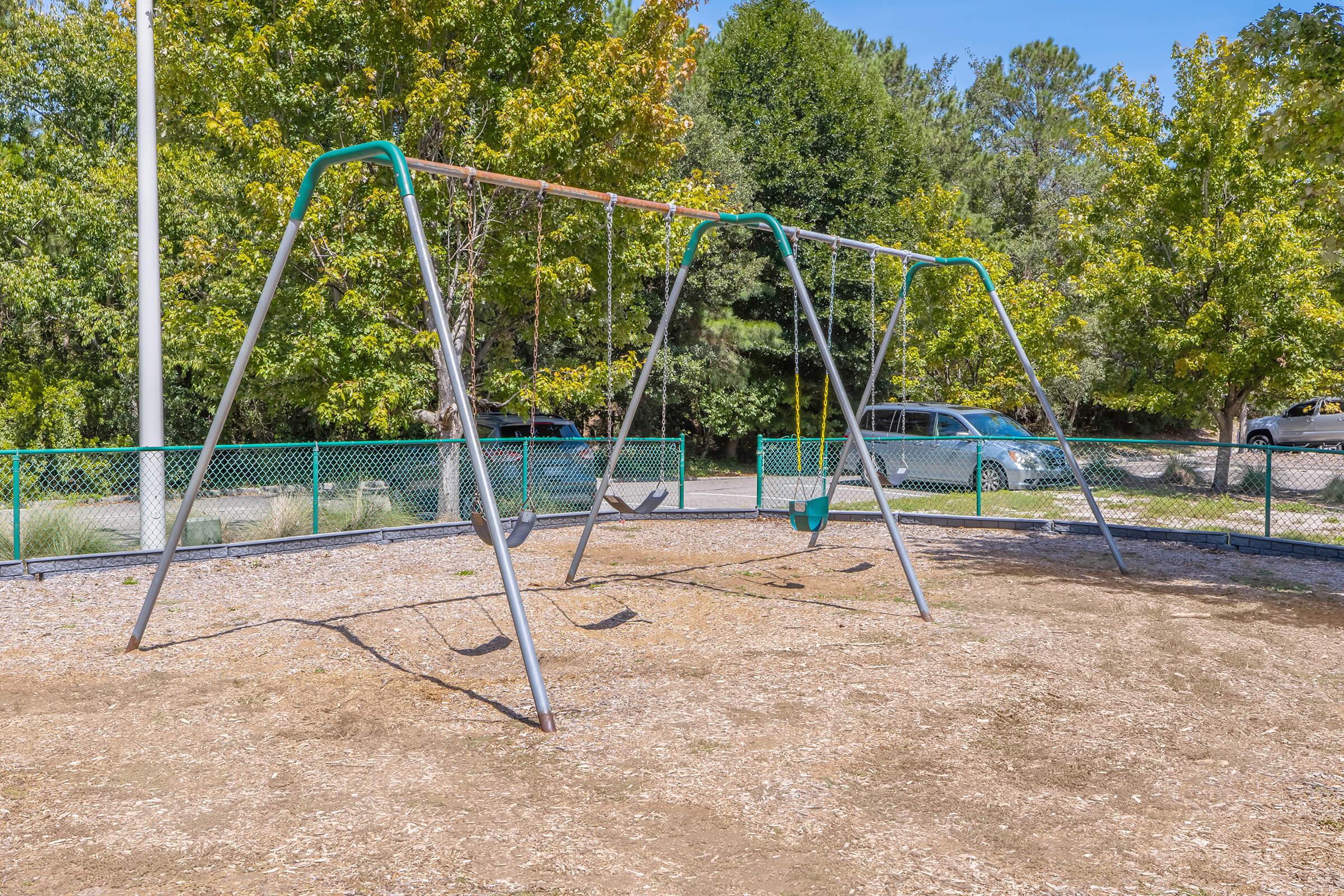 A playground featuring a set of swings, with two swings hanging from a metal frame. The area is surrounded by green fencing and is set against a backdrop of trees and a clear blue sky. Sunlight illuminates the scene, creating a bright and inviting atmosphere for children to play.