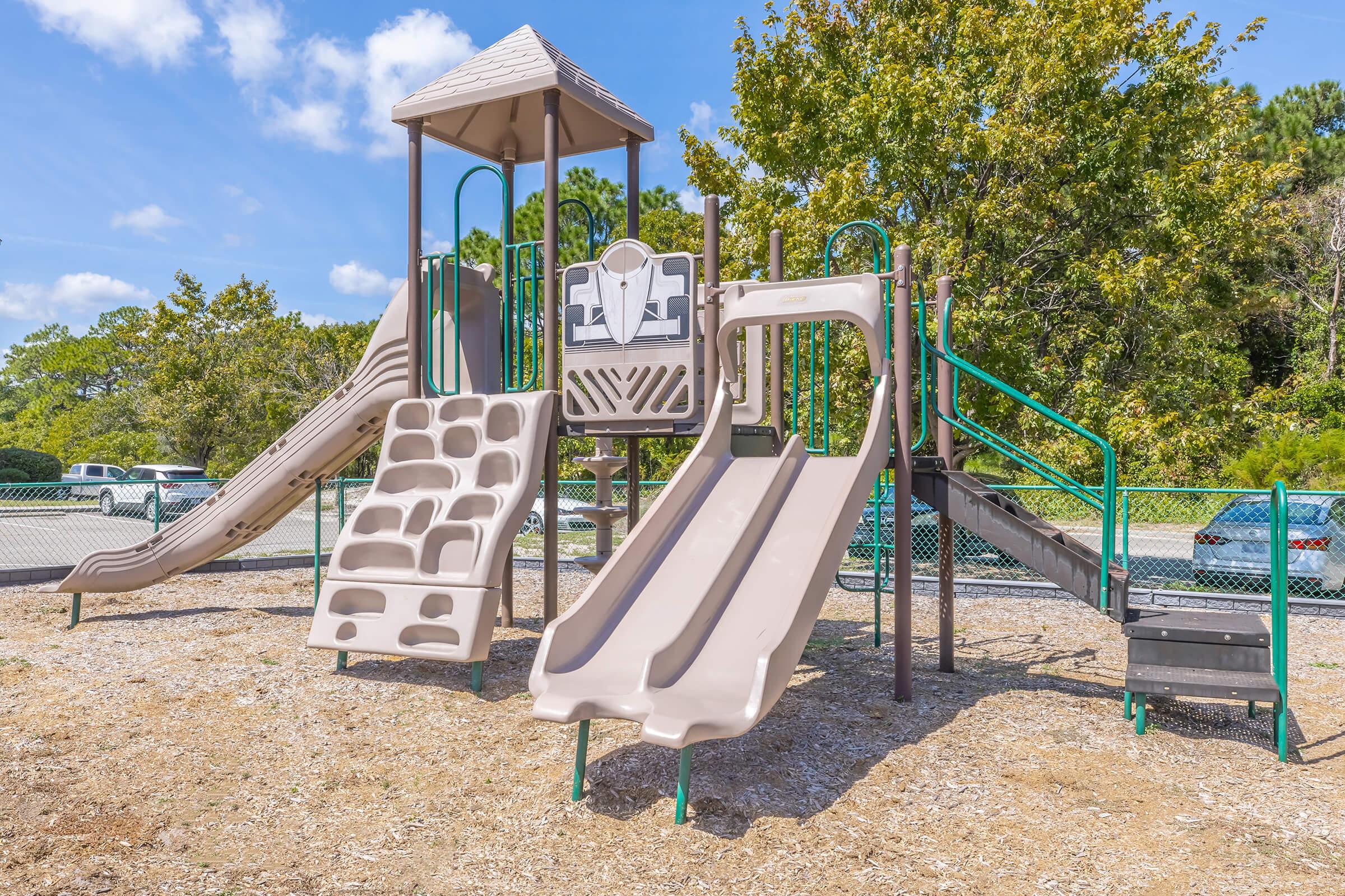 A well-maintained playground featuring a climbing structure with two slides, a climbing wall, and safety rails. The playground is set on a gravel surface, surrounded by green grass, trees, and a fence, under a clear blue sky. It provides a space for children to play and explore.