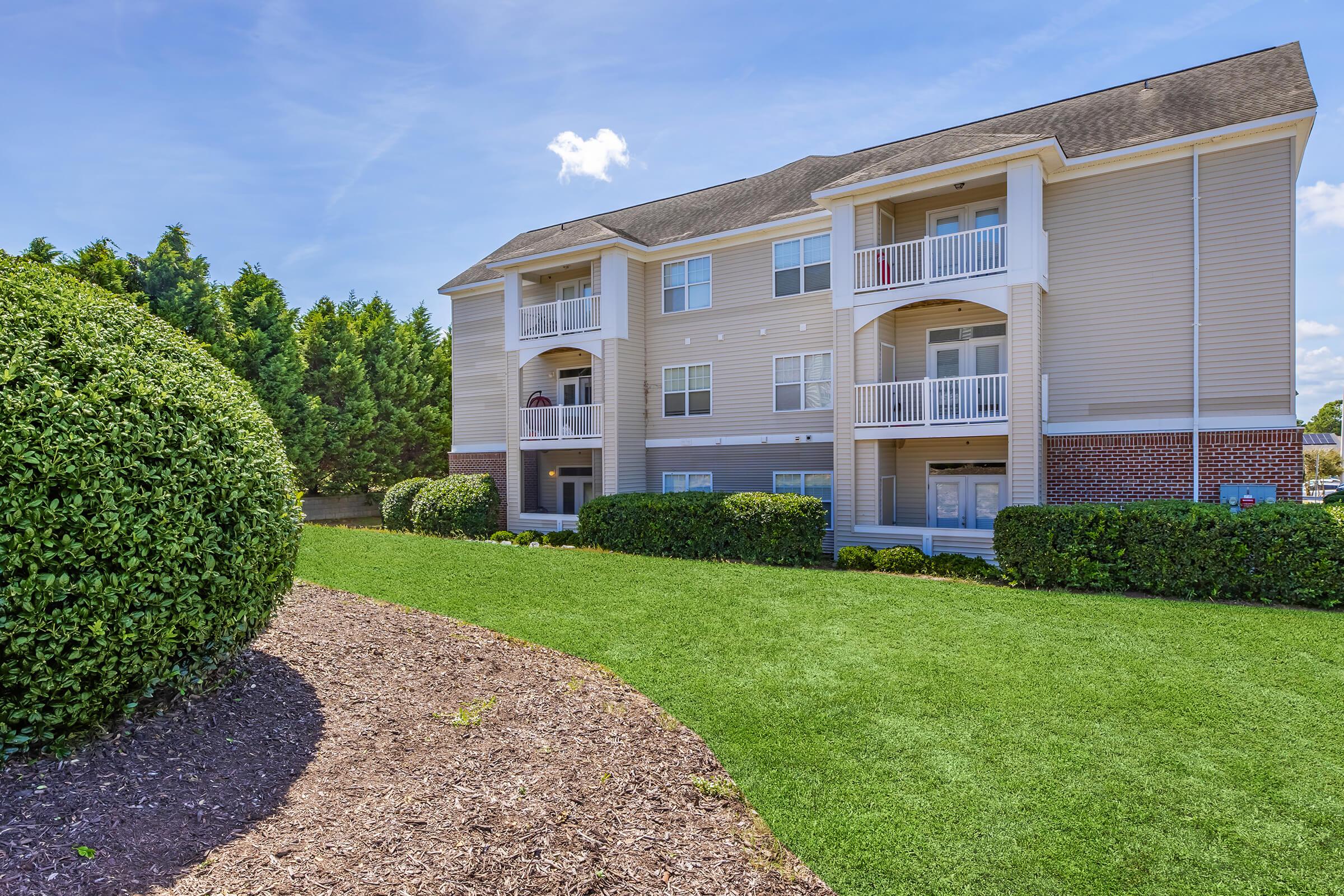 A modern apartment building with three levels, featuring balconies and large windows. The structure is surrounded by well-maintained green lawns and shrubs. In the foreground, a curved garden bed with mulch complements the landscaped area under a clear blue sky with a few white clouds.