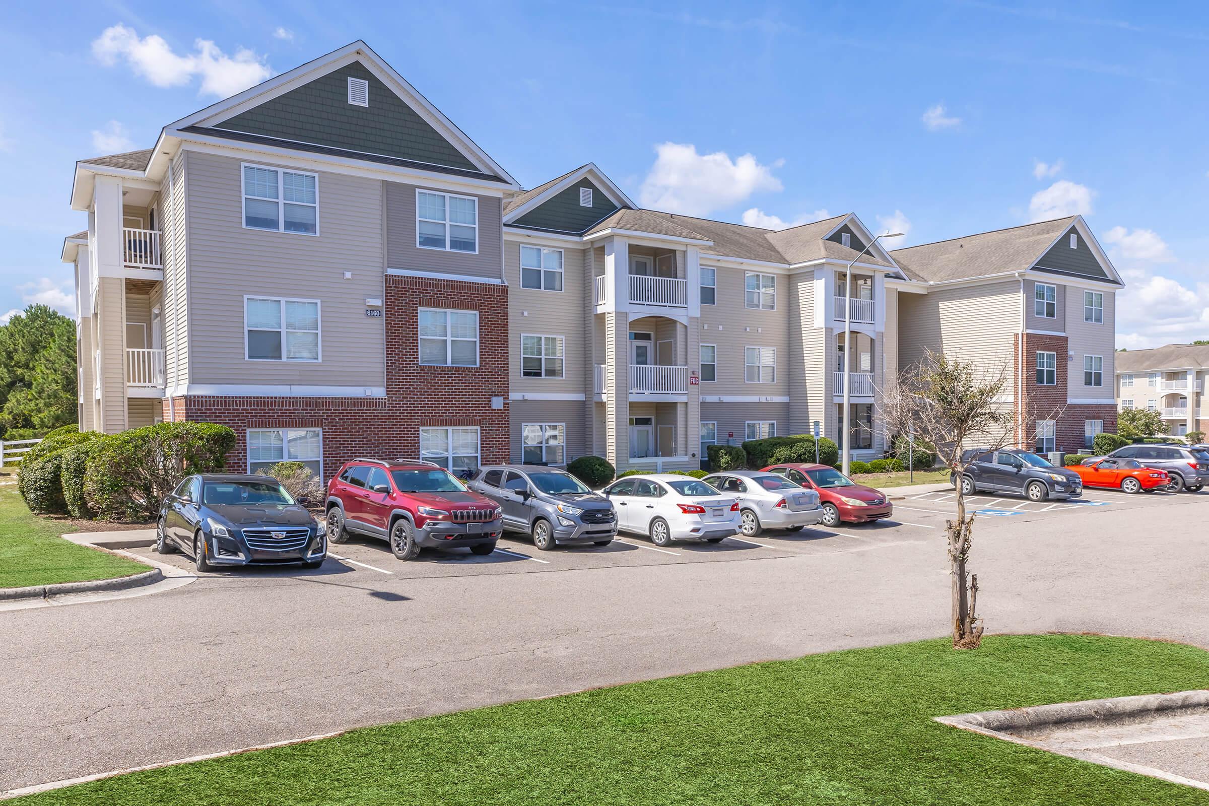 A modern apartment complex featuring three stories with a mix of brick and vinyl siding. A spacious parking lot in front is filled with various vehicles, including sedans and SUVs. Landscaping includes manicured grass and small shrubs, set against a clear blue sky.