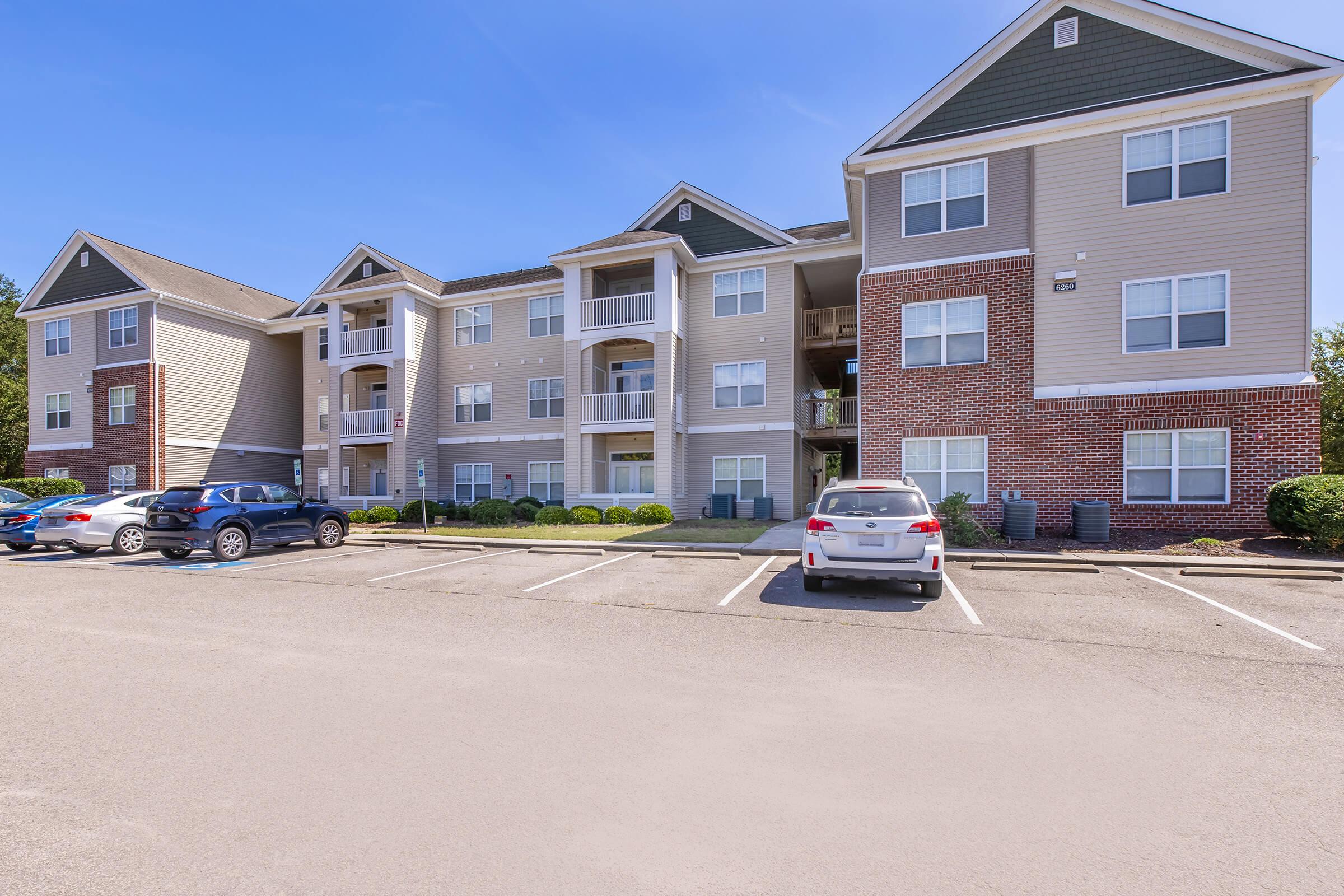 A view of a three-story apartment building with brick and siding exterior, featuring balconies on the upper floors. The foreground shows a parking lot with several parked cars and landscaped greenery surrounding the building, under a clear blue sky.