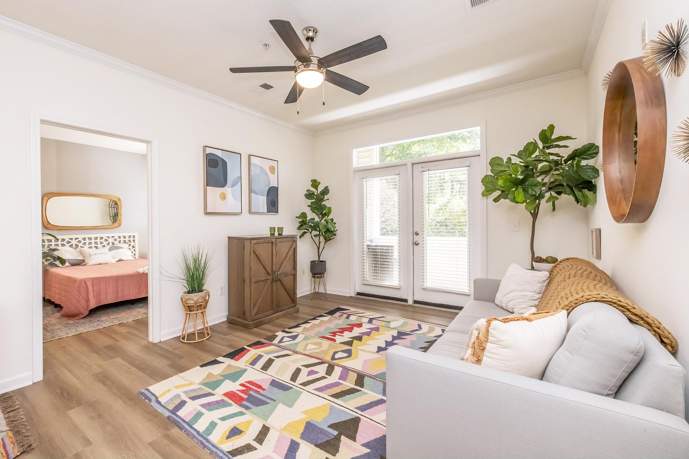 A bright and modern living room featuring a light gray sofa, a colorful geometric area rug, and a ceiling fan. To the left, there's a doorway leading to a bedroom with a cozy bed. Large windows allow natural light in, and potted plants add a touch of greenery to the space.