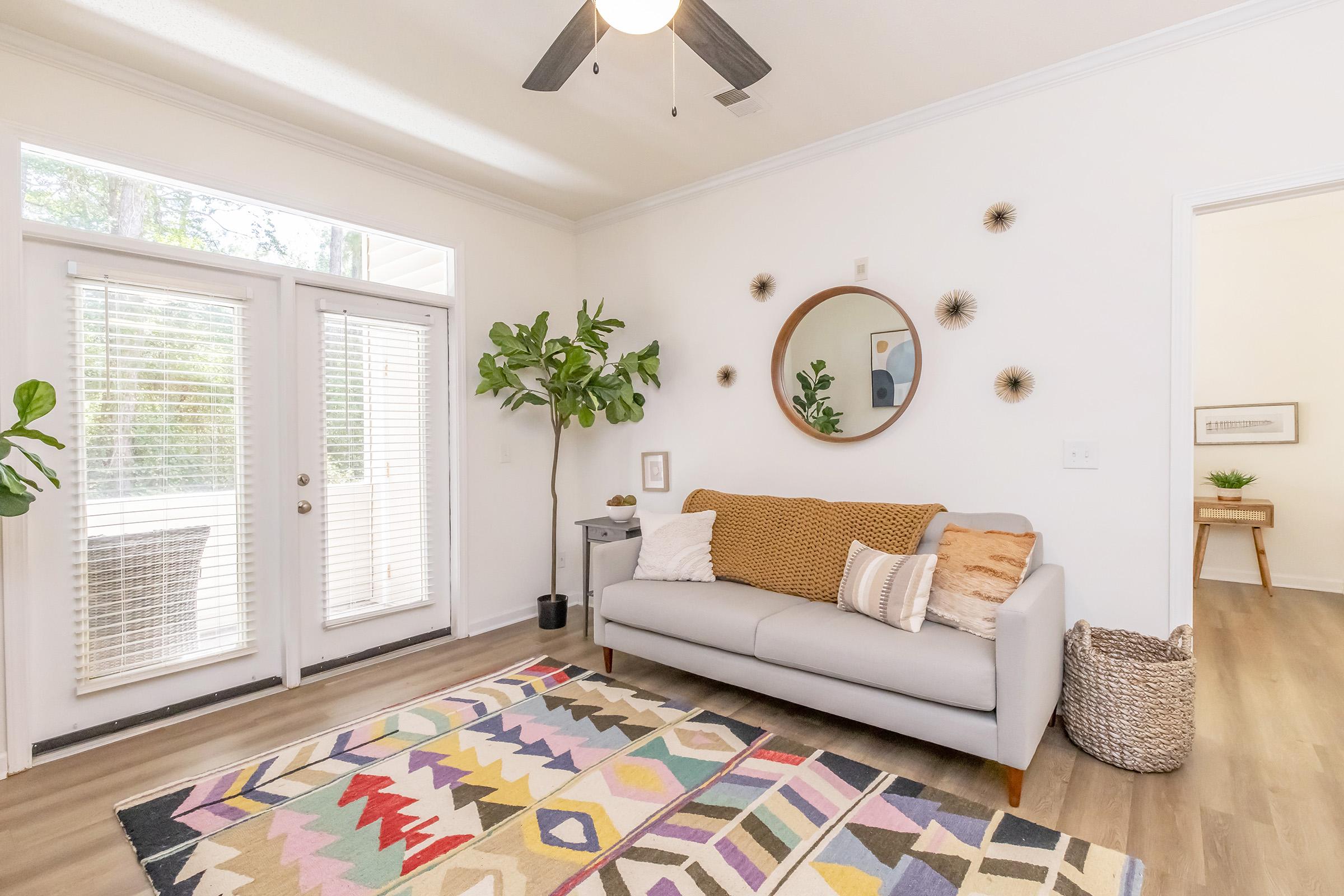 A cozy living room featuring a light gray sofa adorned with decorative pillows, a colorful geometric area rug, and a round mirror on the wall. Large windows with blinds allow natural light, complemented by indoor plants and minimalistic decor. The space feels inviting and bright.