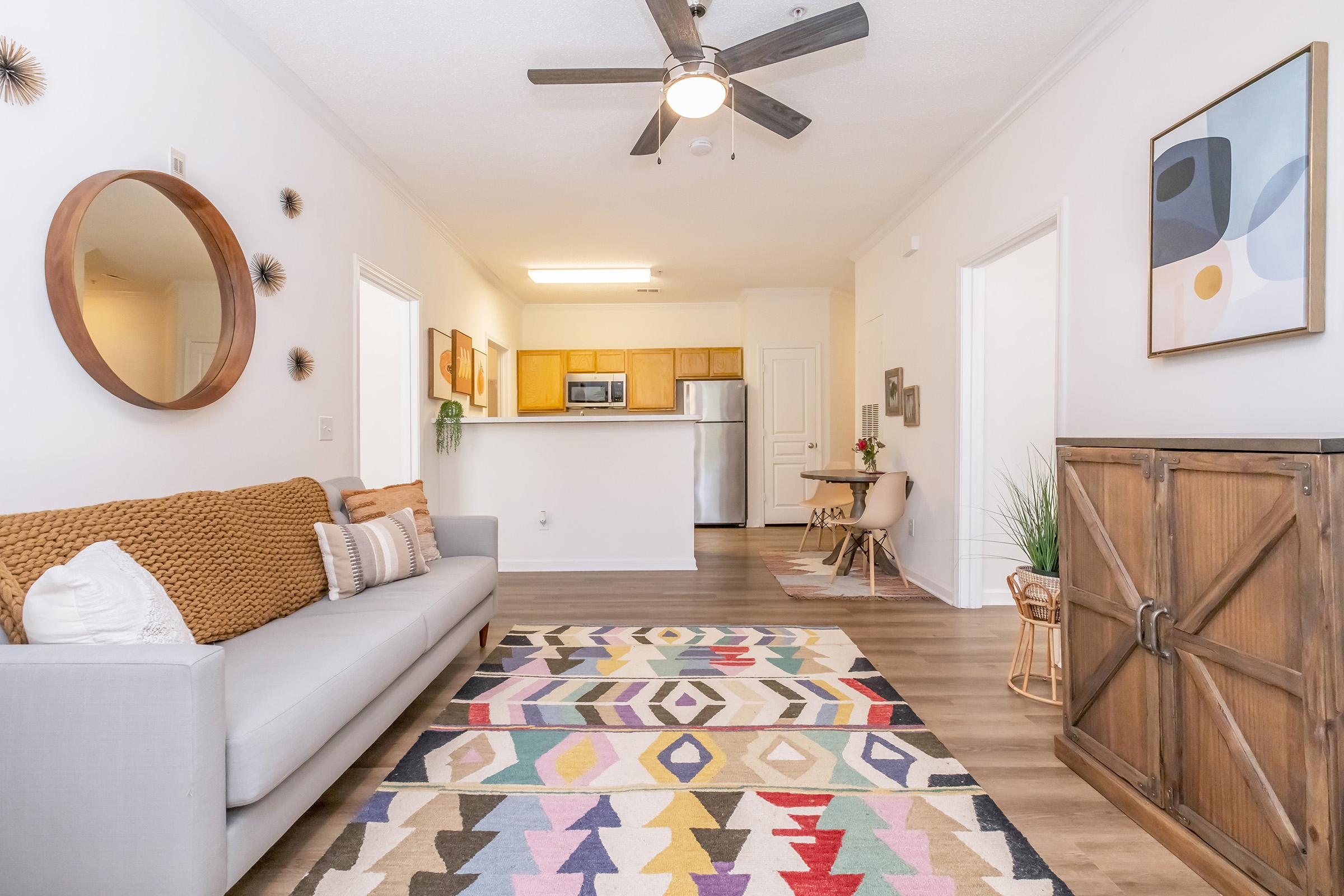 A cozy living room featuring a gray sofa with decorative pillows, a colorful geometric rug, a round mirror, and a wooden accent cabinet. The space has an open layout leading to a kitchen area with light wood cabinets and stainless steel appliances. Natural light illuminates the room, creating a welcoming atmosphere.