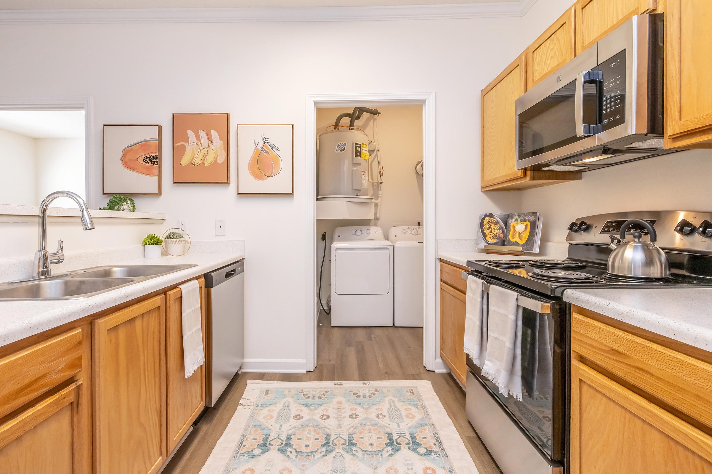 A modern kitchen featuring wooden cabinets, stainless steel appliances, and a double sink. The space includes a decorative rug and framed artwork on the wall. In the background, there is a laundry area with a washer and dryer. Bright and airy with natural light.