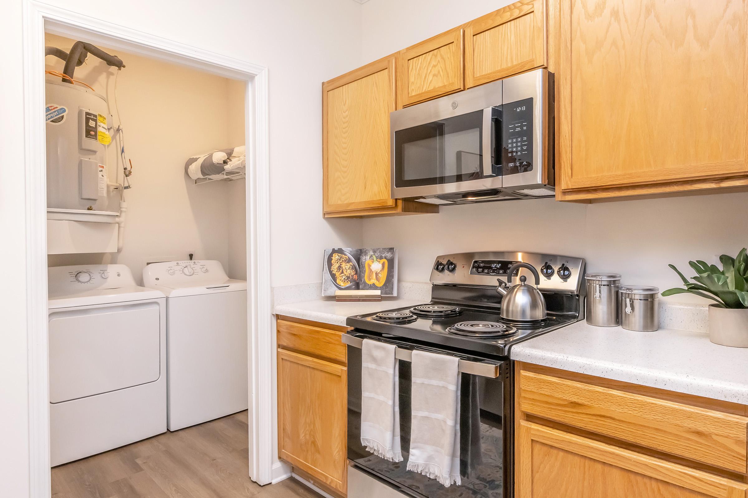 A modern kitchen featuring wooden cabinets, a stainless steel stove, and a microwave above. A washing machine and dryer are visible in a laundry area through an open door. Countertop decor includes a plant and framed artwork, while towels hang from the oven. The space is clean and well-lit, showcasing a functional layout.