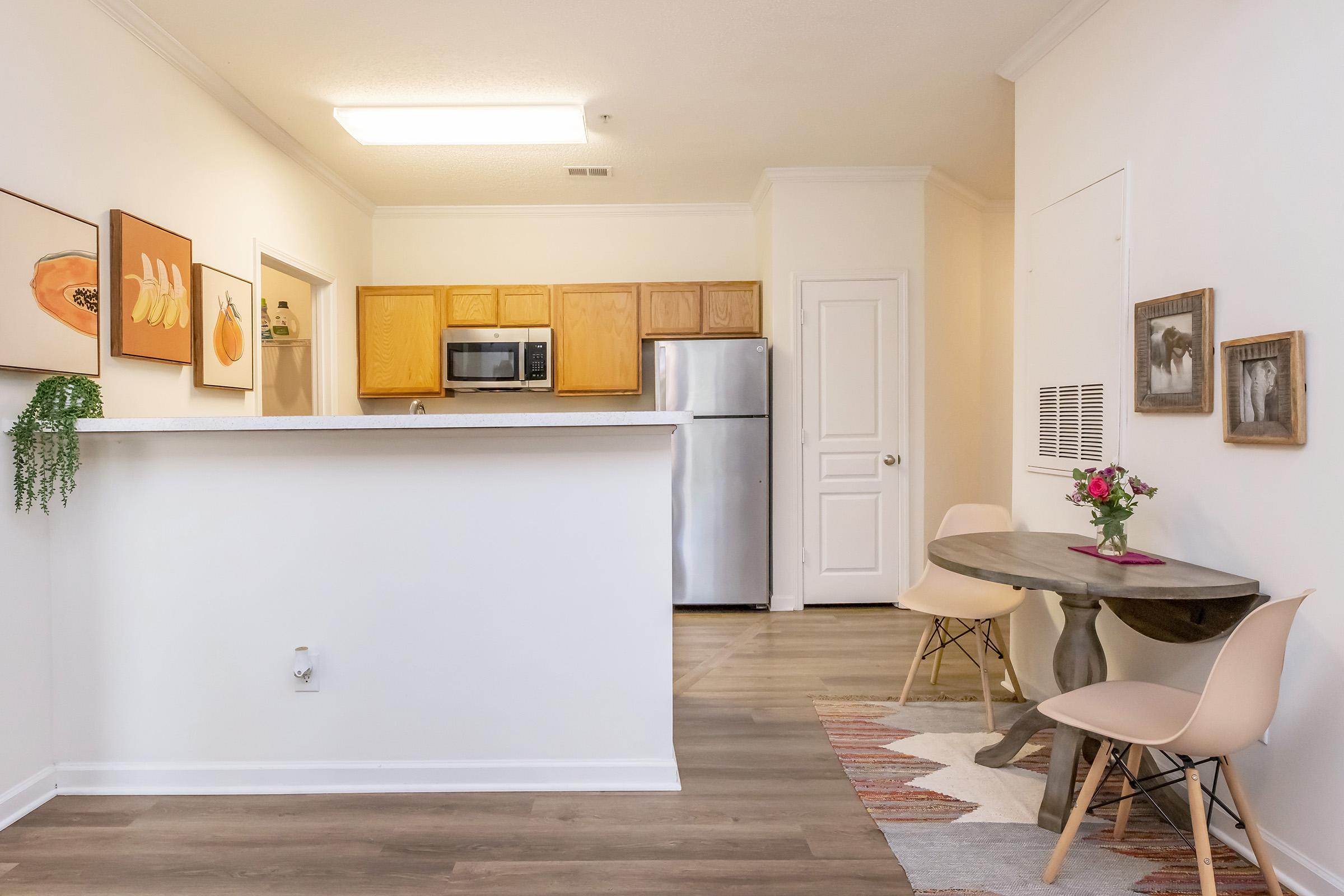 A bright and inviting apartment interior featuring a small dining area with a round table and two chairs. The kitchen is visible in the background with wooden cabinets and stainless steel appliances. Wall art adorns the space, adding a pop of color, while a vase with flowers sits on the table. The flooring is light wood.