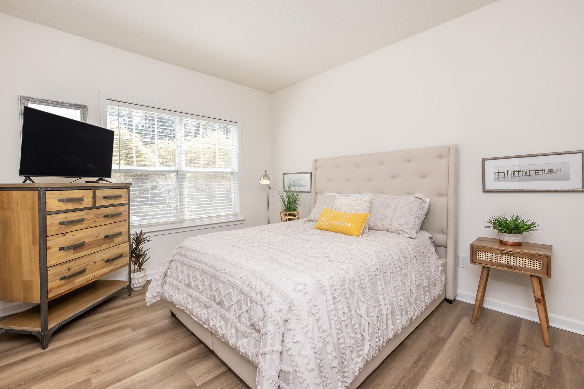 A cozy bedroom featuring a light-colored bed with a textured white bedspread and a yellow decorative pillow. A small nightstand with a plant sits beside the bed. A wooden dresser with a TV on top is positioned against one wall, and a bright window allows natural light to fill the space, enhancing the inviting atmosphere.