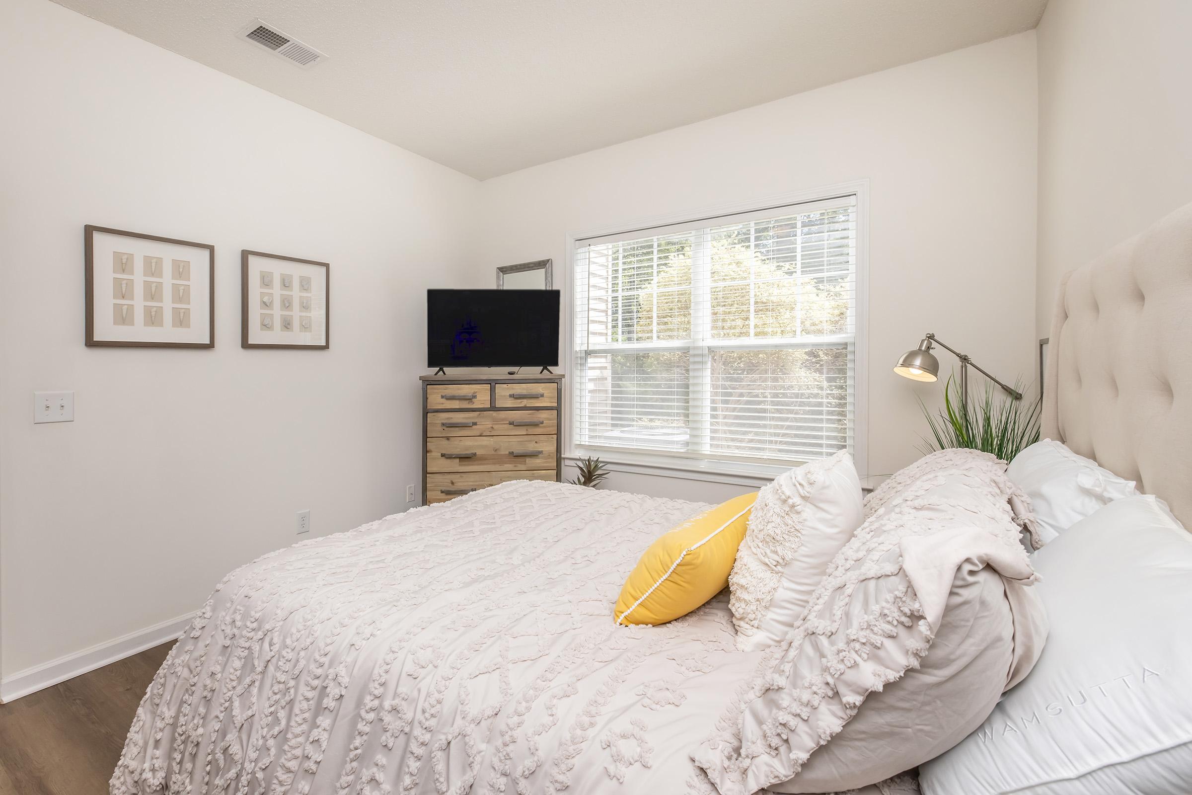 A cozy bedroom featuring a light-colored bed with textured bedding and a yellow accent pillow. There’s a wooden dresser with a television on top, a table lamp, and a small plant by the window. Natural light fills the room, highlighting the neutral decor and minimalist style.
