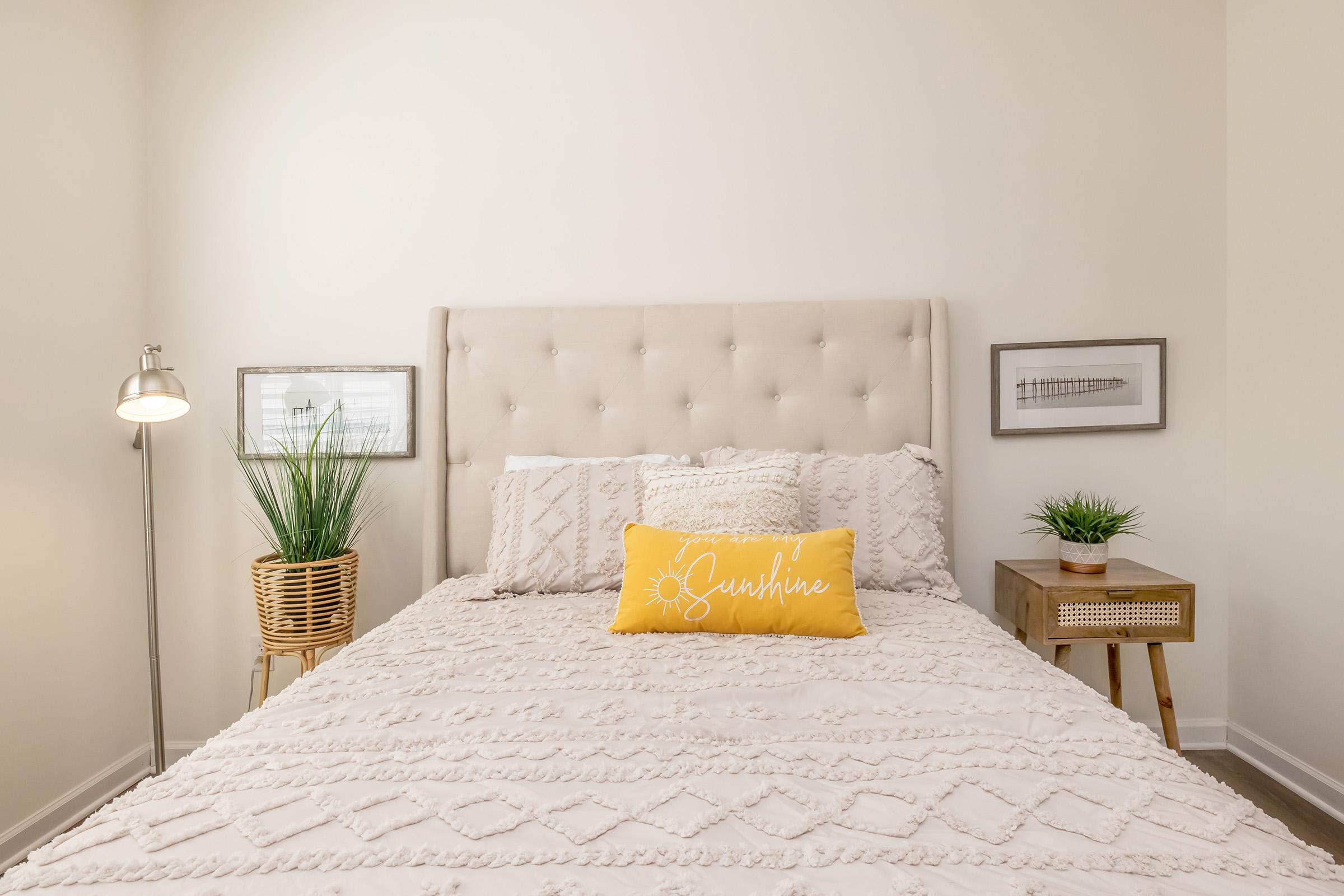 Cozy bedroom featuring a beige tufted headboard, a neatly made bed with a textured white blanket, and a yellow "Sunshine" pillow. A small nightstand with plants and a lamp is beside the bed, and framed artwork is displayed on the wall, enhancing the warm, inviting atmosphere.