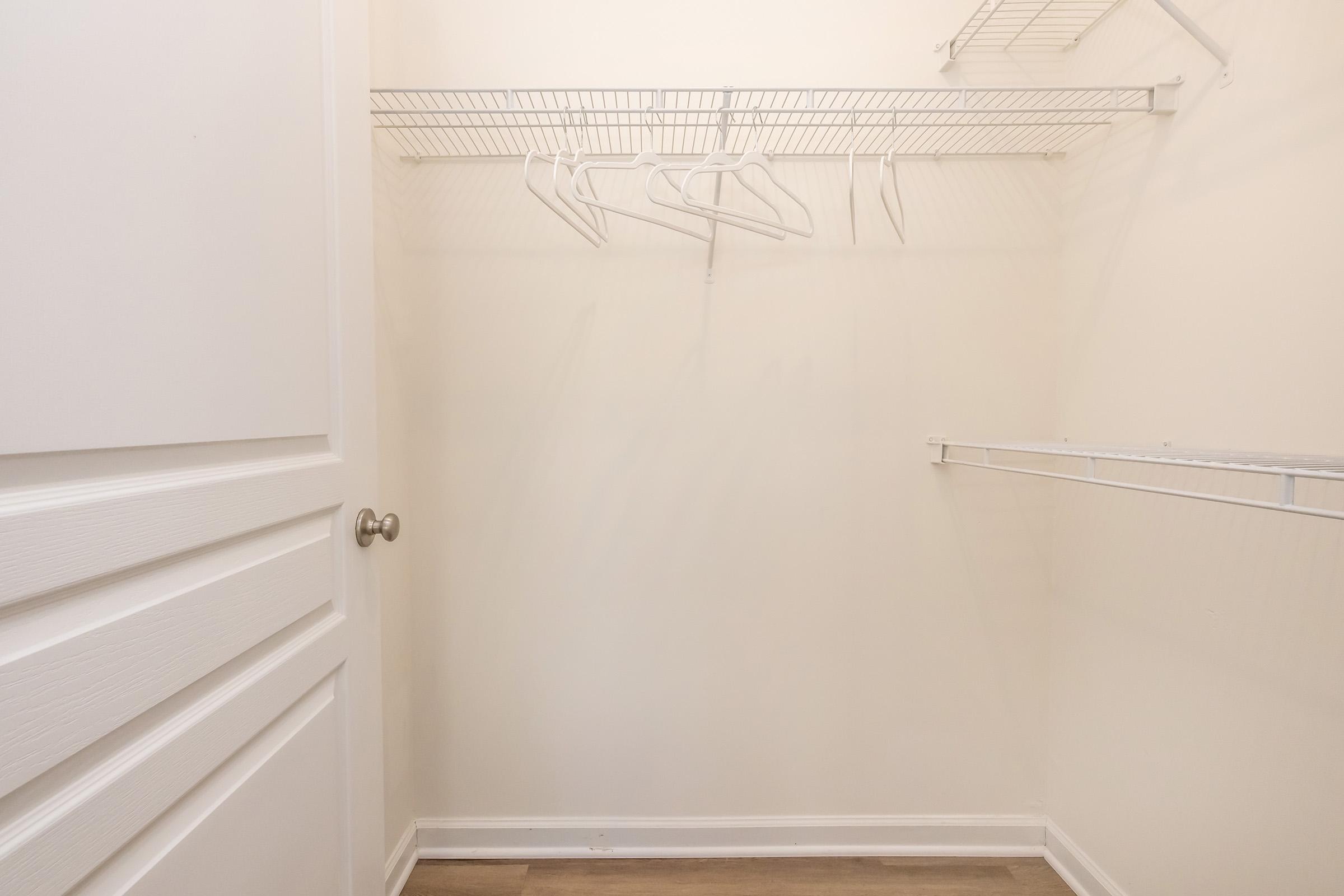 A spacious, empty closet with white walls and shelves. There are a few white hangers hanging from a wire rack. The closet features a closed door with a silver knob, and the floor is covered in light-colored flooring. The space appears clean and organized.