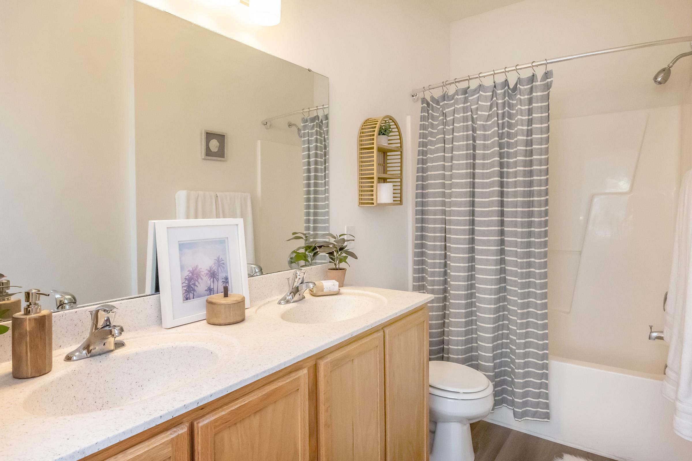 A modern bathroom featuring a double vanity with two sinks and wooden cabinets. A large mirror above the vanity reflects soft light. There's a shower area with a gray striped curtain, a white toilet, and decorative elements like plants and framed artwork. The overall color scheme is neutral and calming.