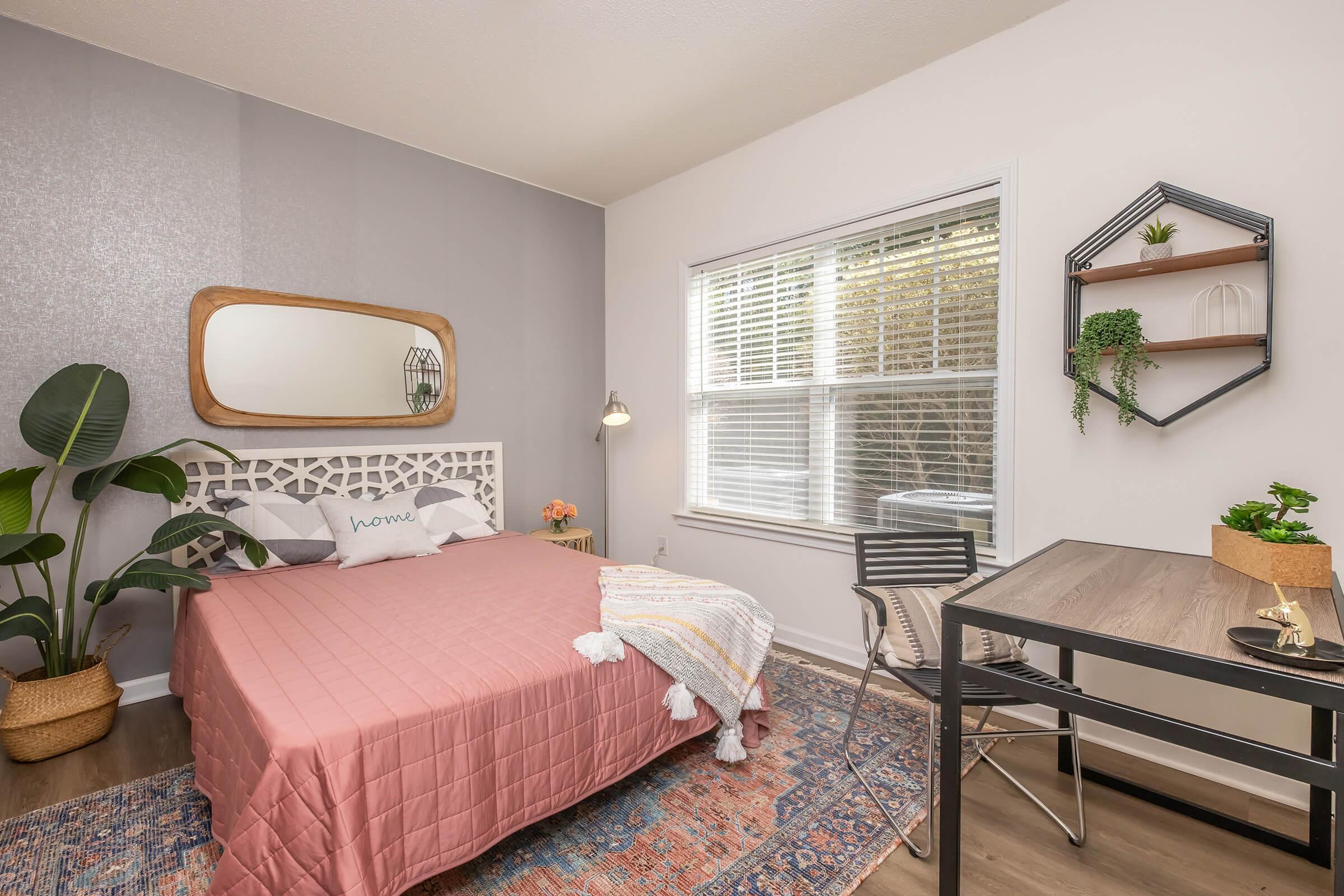 A cozy bedroom featuring a pink quilted bedspread with decorative pillows, a large mirror above the bed, and a small desk with a chair. Soft lighting highlights the room's greenery, including a large plant, and decorative shelves with plants and accessories are mounted on the wall. The overall decor is modern and inviting.