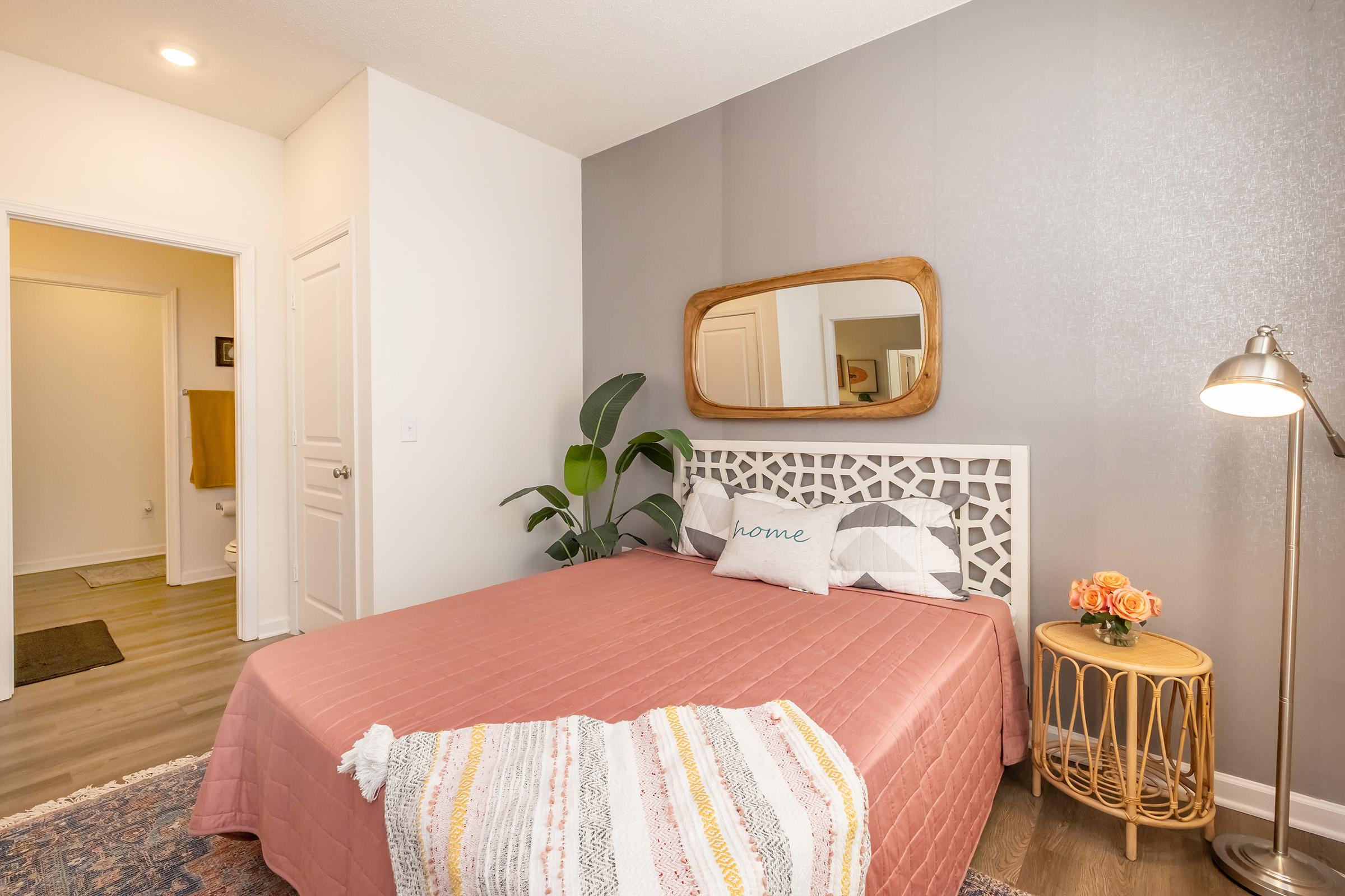 A cozy bedroom featuring a pink bedspread, decorative pillows, and a wooden-framed mirror. There are potted plants, a stylish bedside table with flowers, and a warm, inviting wall color. The room has a soft, modern aesthetic with light hardwood flooring and a doorway leading to another area.