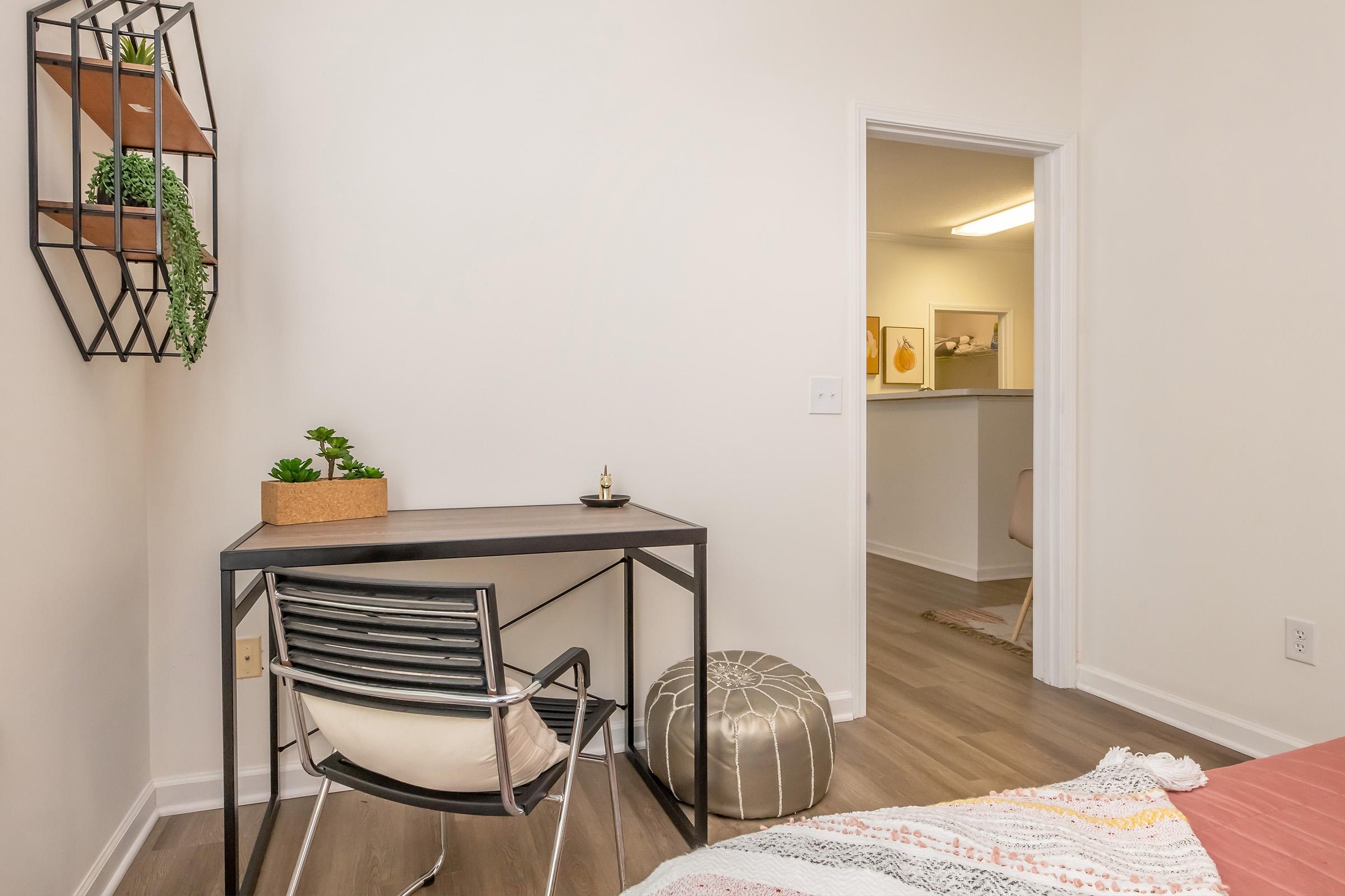 A minimalist room featuring a small desk with a chair, decorated with a potted plant. A geometric wall shelf holds another plant. The room has neutral-colored walls and a glimpse of a hallway with light-colored decor in the background. A cozy bed with a soft pink blanket is visible.