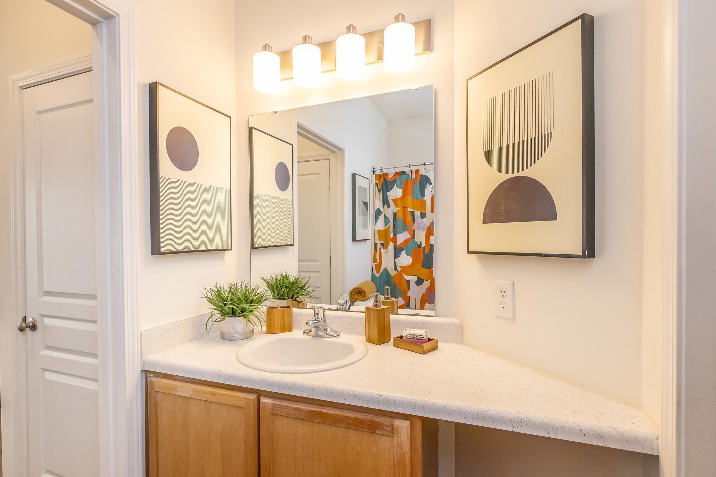 A modern bathroom with a light-colored countertop and sink. The wall features two abstract art pieces, and there’s a colorful shower curtain. Plants and wooden decor items are placed on the counter, enhancing the aesthetic. Bright lighting is provided by four light fixtures above the mirror.
