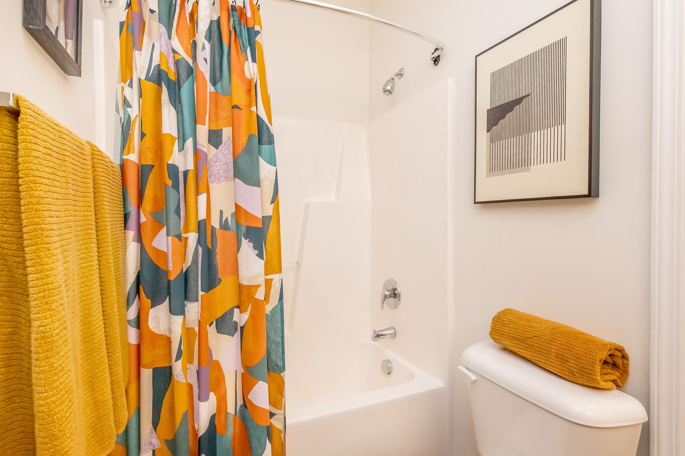 A brightly colored shower curtain with abstract patterns hangs in a bathroom. A yellow towel is neatly placed on a white toilet, and a matching yellow bath mat is visible. A modern art piece is framed on the wall, adding a touch of decor to the space. The overall ambiance is vibrant and cheerful.