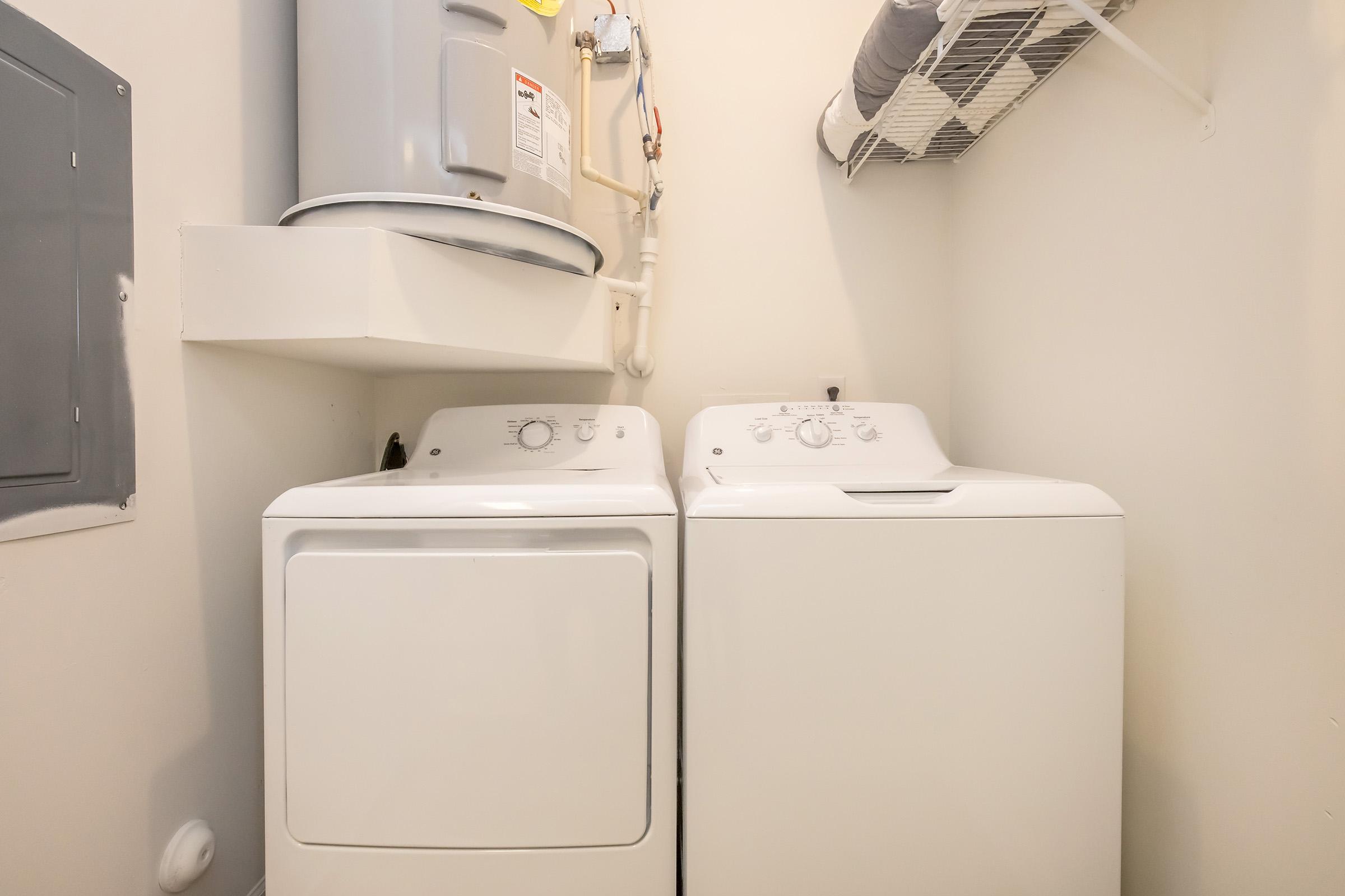 A clean laundry room featuring a stacked washer and dryer set, with a water heater mounted on the wall above. There is a metal shelf above the appliances, and the walls are painted a neutral color, providing a tidy and organized appearance.