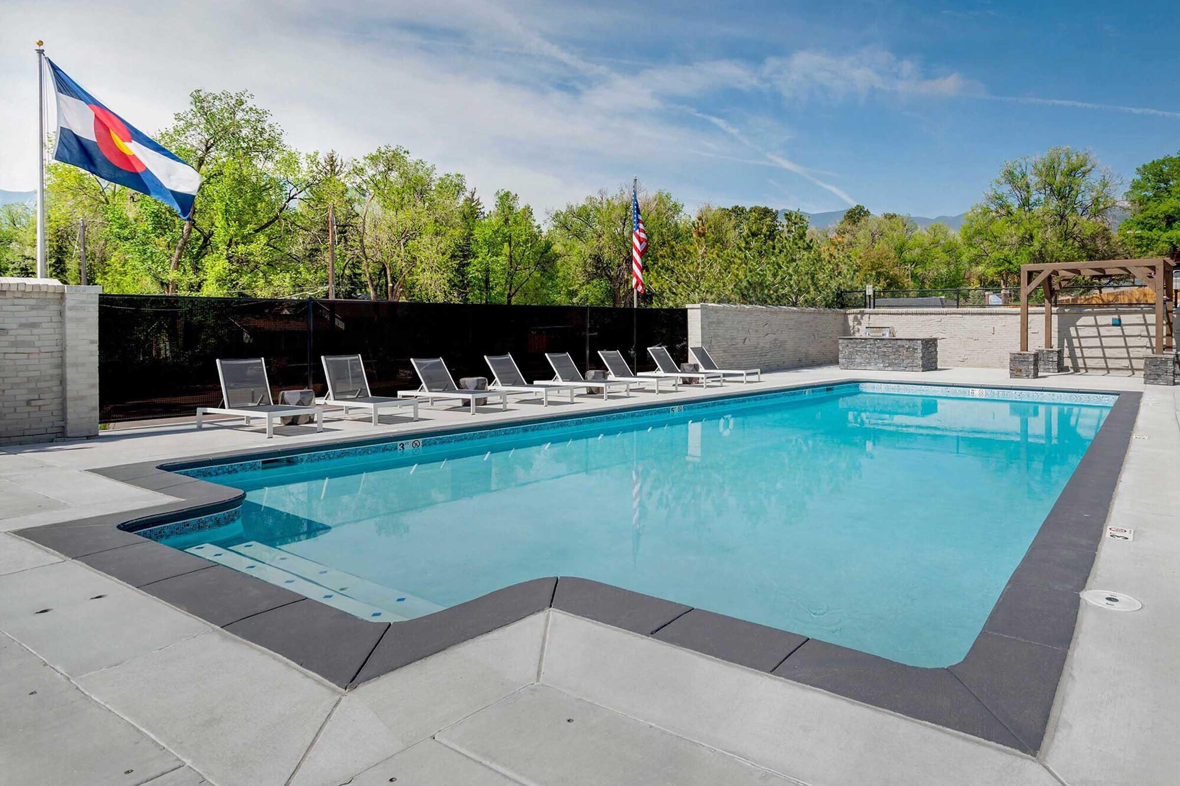 Outdoor swimming pool with clear blue water, surrounded by lounge chairs. Colorado state flag and American flag waving in the background. Lush green trees and a bright blue sky add to the serene atmosphere of the area. A modern design with a sleek pool edge.