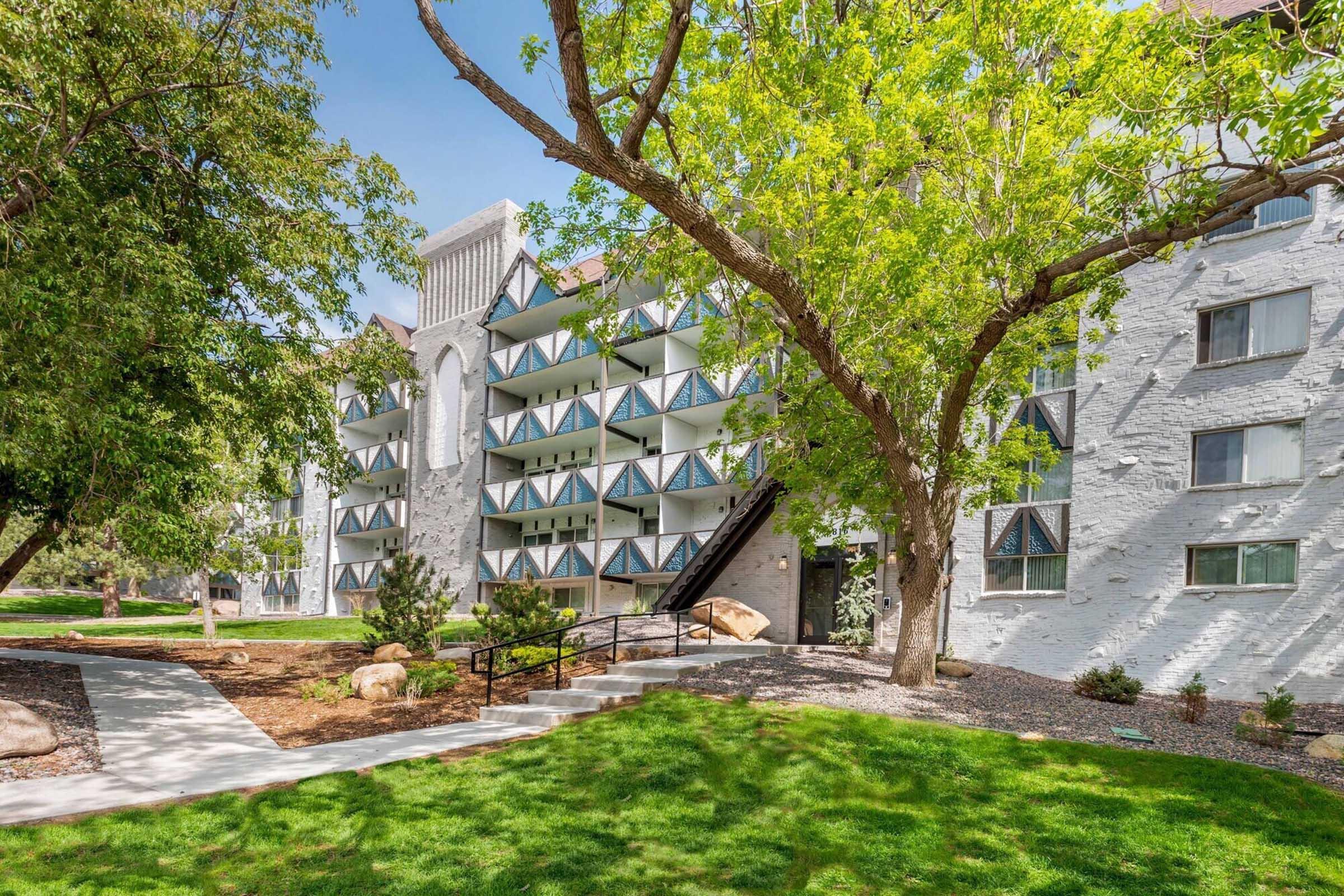 A multi-story white building with balconies surrounded by green grass and trees. A pathway leads to the entrance, and the landscaping features rocks and shrubs. The sky is clear and blue, enhancing the inviting atmosphere of the property.
