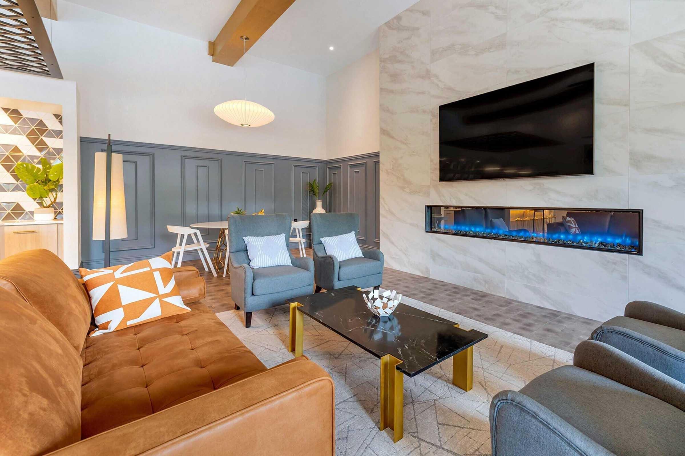 Modern living room featuring a brown leather sofa with a geometric orange and white pillow, two blue armchairs, a glass coffee table with decorative items, and a sleek fireplace beneath a large wall-mounted TV. The walls are adorned with a mix of textures, and there’s a dining area in the background.