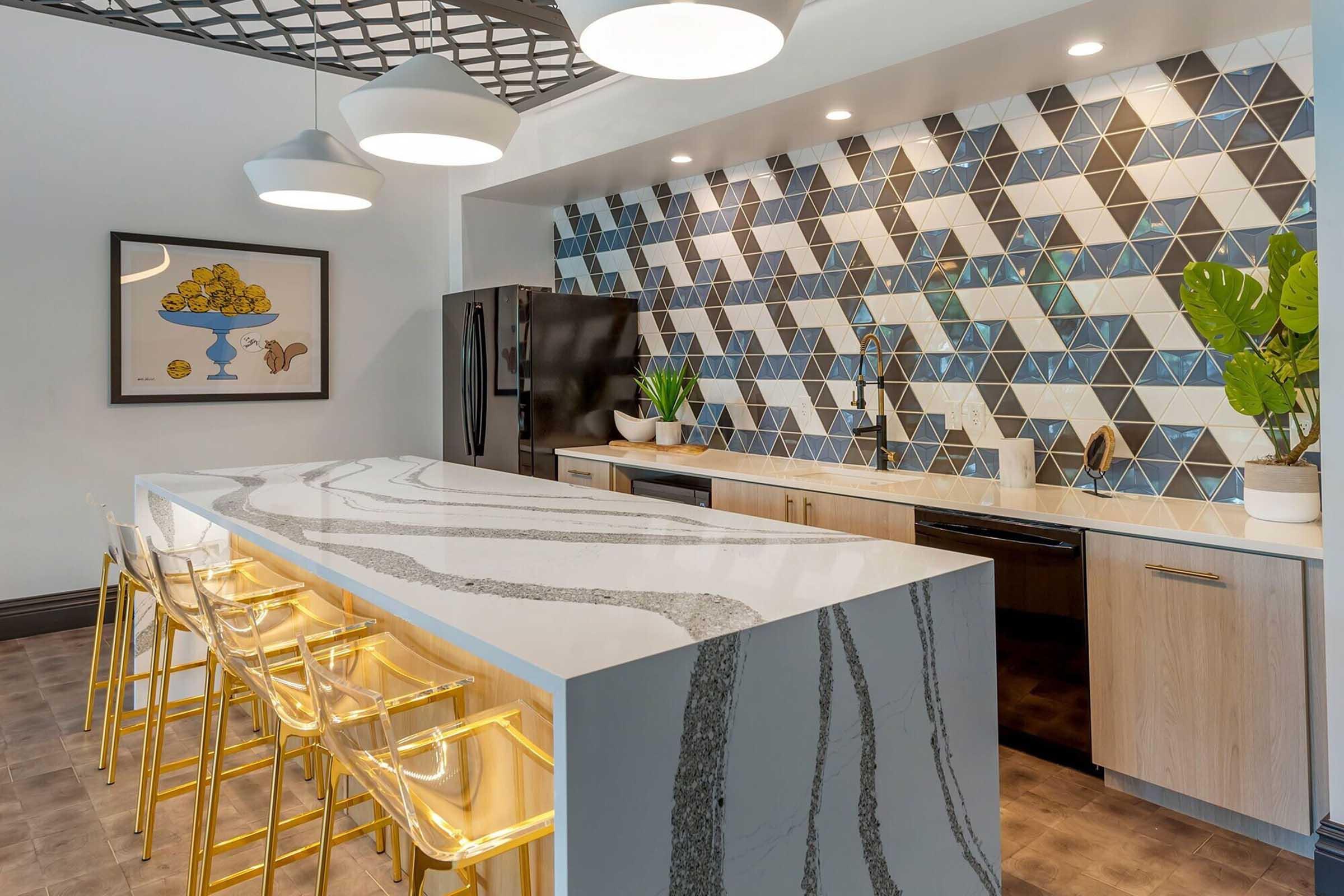 Modern kitchen featuring a stylish island with a marbled countertop and gold-accented clear chairs. The backsplash features a geometric tile design in blue and white tones. A large black refrigerator and sleek appliances complete the contemporary look, while potted plants add a touch of greenery.