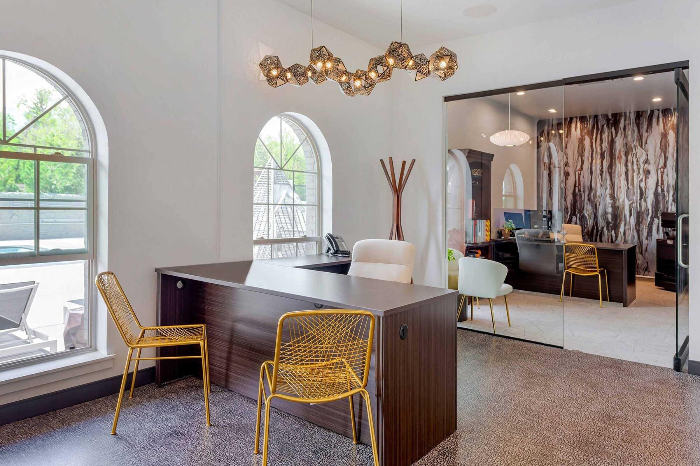Modern office interior featuring a dark wood reception desk, yellow wire chairs, and a unique chandelier. Large arched windows allow natural light to fill the space, and a glass partition reflects a stylish work area in the background. Subtle textures and colors create a warm, inviting atmosphere.