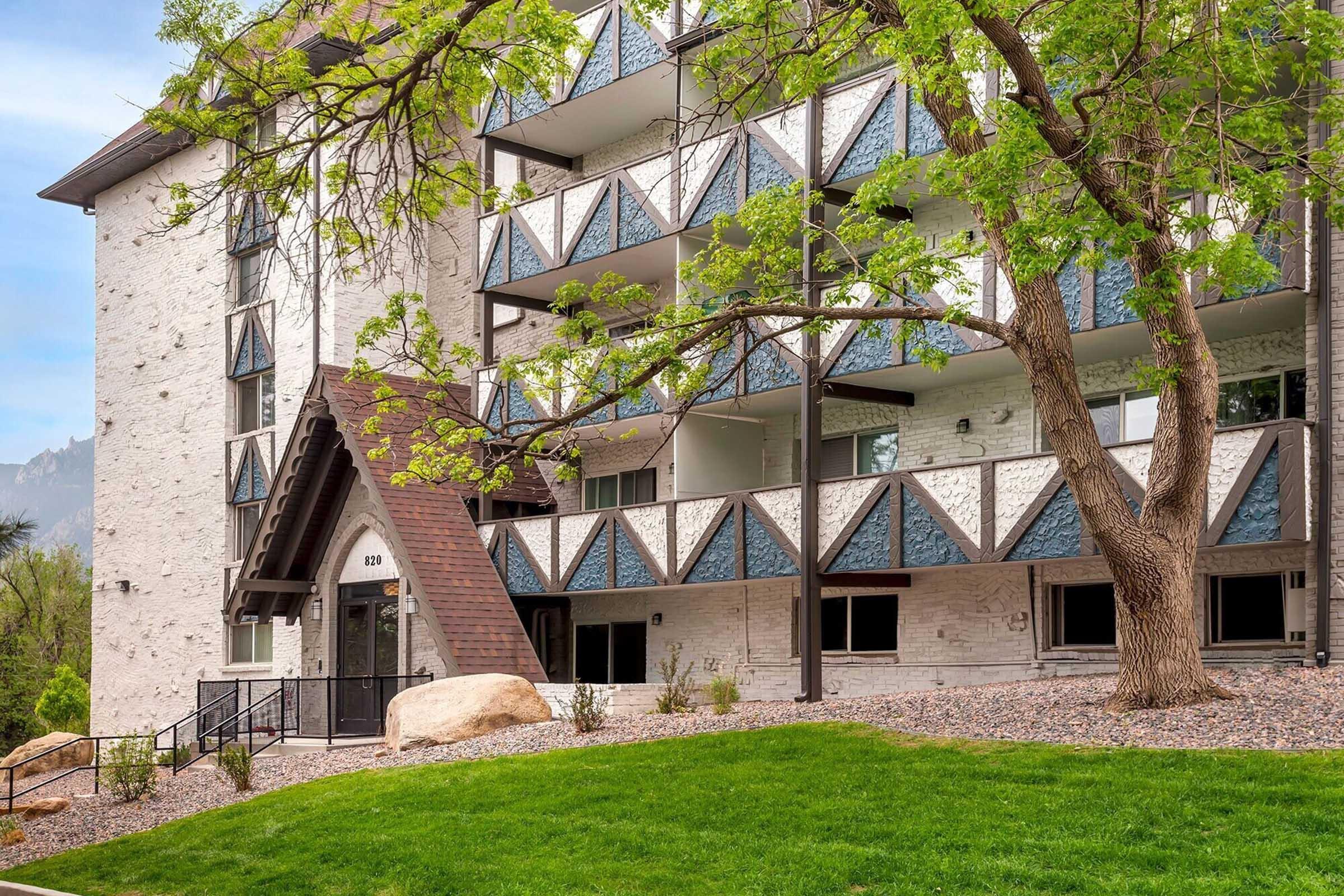A multi-story building featuring a white exterior with decorative blue accents on the balconies. In front, there is a landscaped area with green grass and a tree. The entrance has a peaked roof and steps leading up to it, surrounded by rocks and shrubs.
