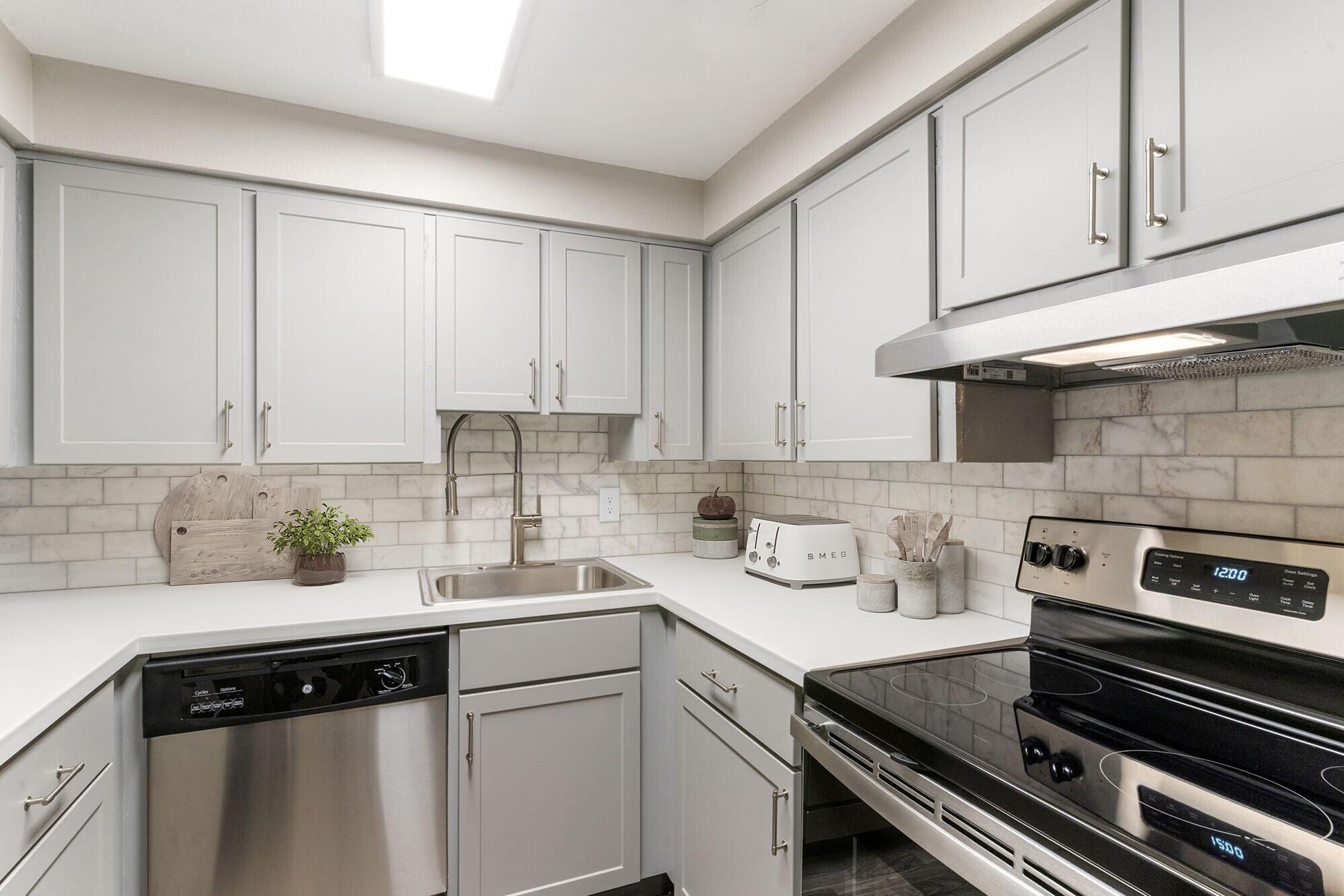 A modern kitchen featuring gray cabinets, white countertops, and a stainless steel sink. The kitchen includes a dishwasher, an electric stove, and a white-tiled backsplash. Light from a ceiling fixture brightens the space, which is decorated with small plants and kitchen appliances.