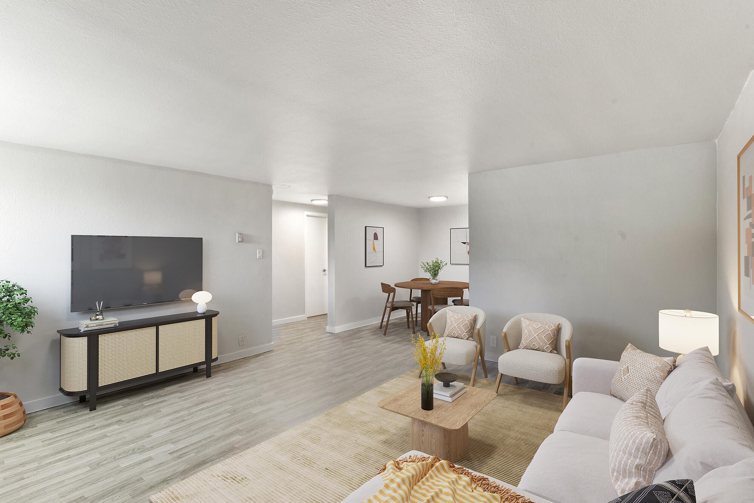 A modern living room with light gray walls and wood laminate flooring. It features a large flat-screen TV on a stylish entertainment center, a beige sectional sofa, a coffee table with decorative items, a dining area with a wooden table and chairs, and a potted plant adding a touch of greenery.