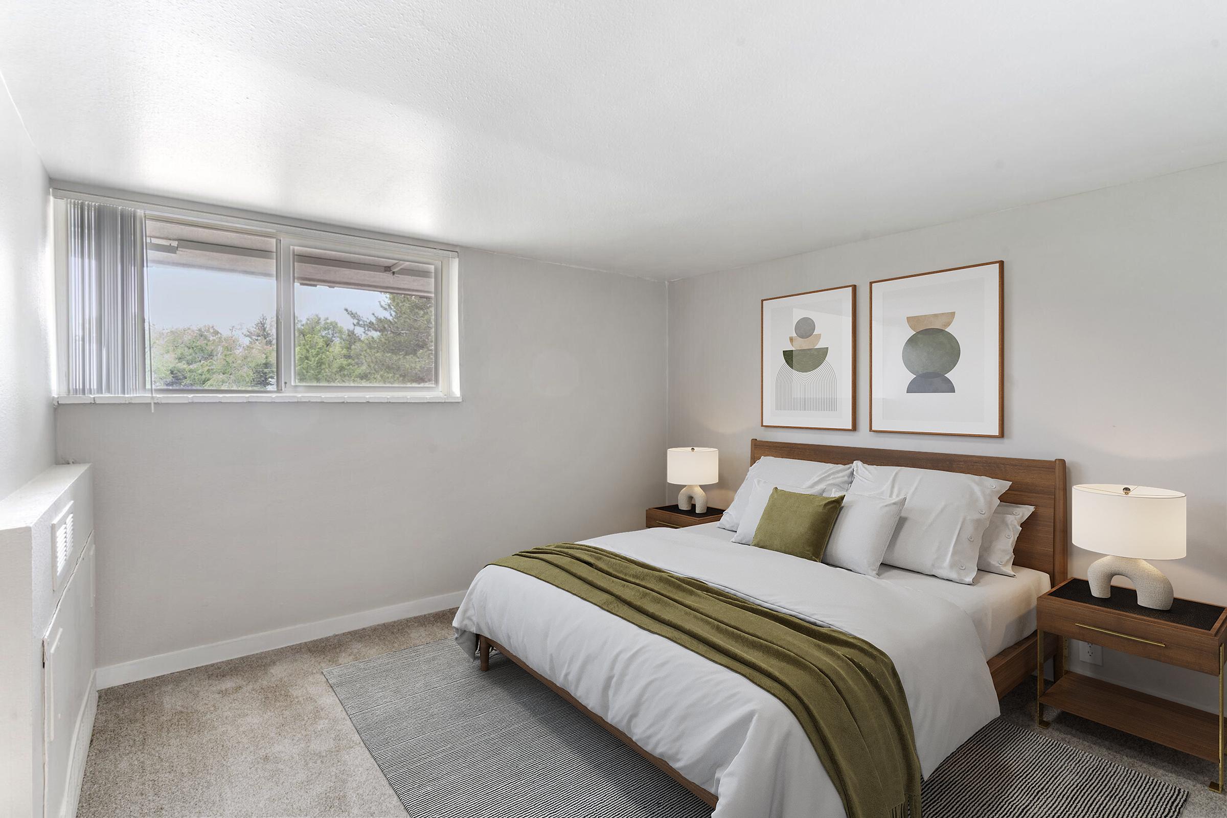 A stylish, modern bedroom featuring a large bed with white linens and a green throw. There are two framed abstract art pieces on the wall, bedside tables with lamps, and a window allowing natural light to enter. The room has a neutral color scheme and a cozy carpeted floor.