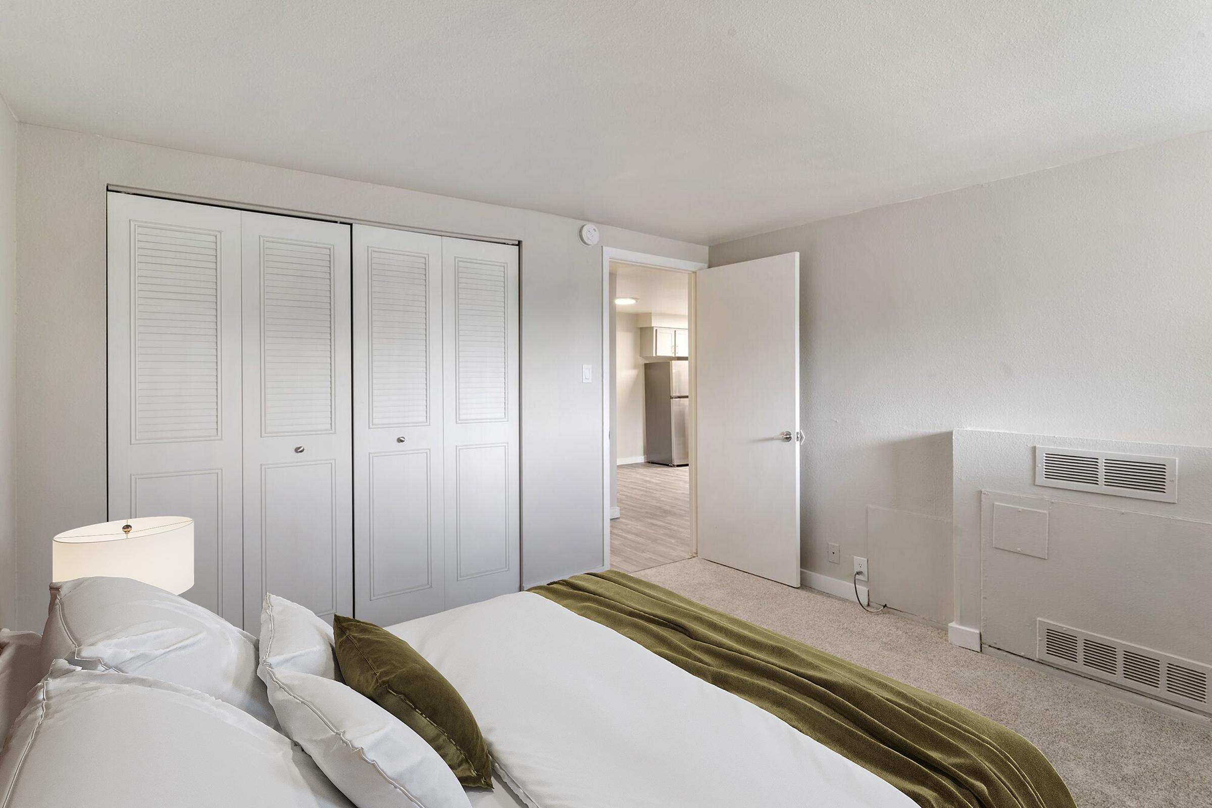 A cozy bedroom featuring a neatly made bed with white sheets and a green throw. There is a bedside lamp beside the bed, and double closet doors are visible. A doorway leads to another room, hinting at an open layout. The walls are painted in neutral tones, creating a calm atmosphere.