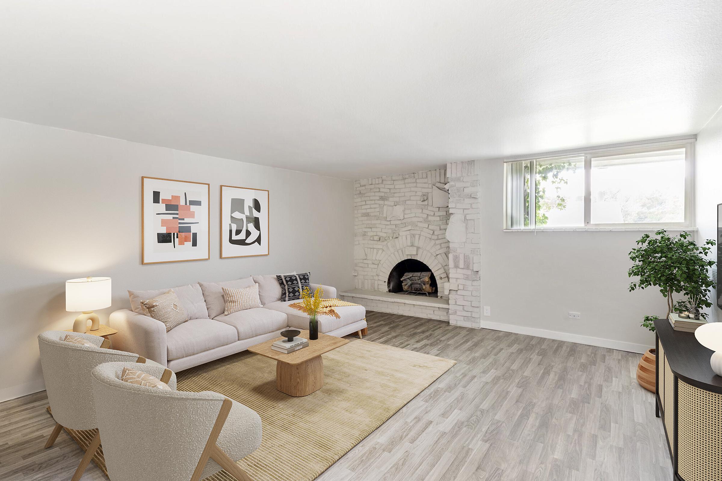 A modern living room featuring a light-colored sofa with decorative pillows, two armchairs, a wooden coffee table, and a cozy stone fireplace. The walls are adorned with abstract art, and a large window allows natural light to fill the space. A plant and a TV are also present.
