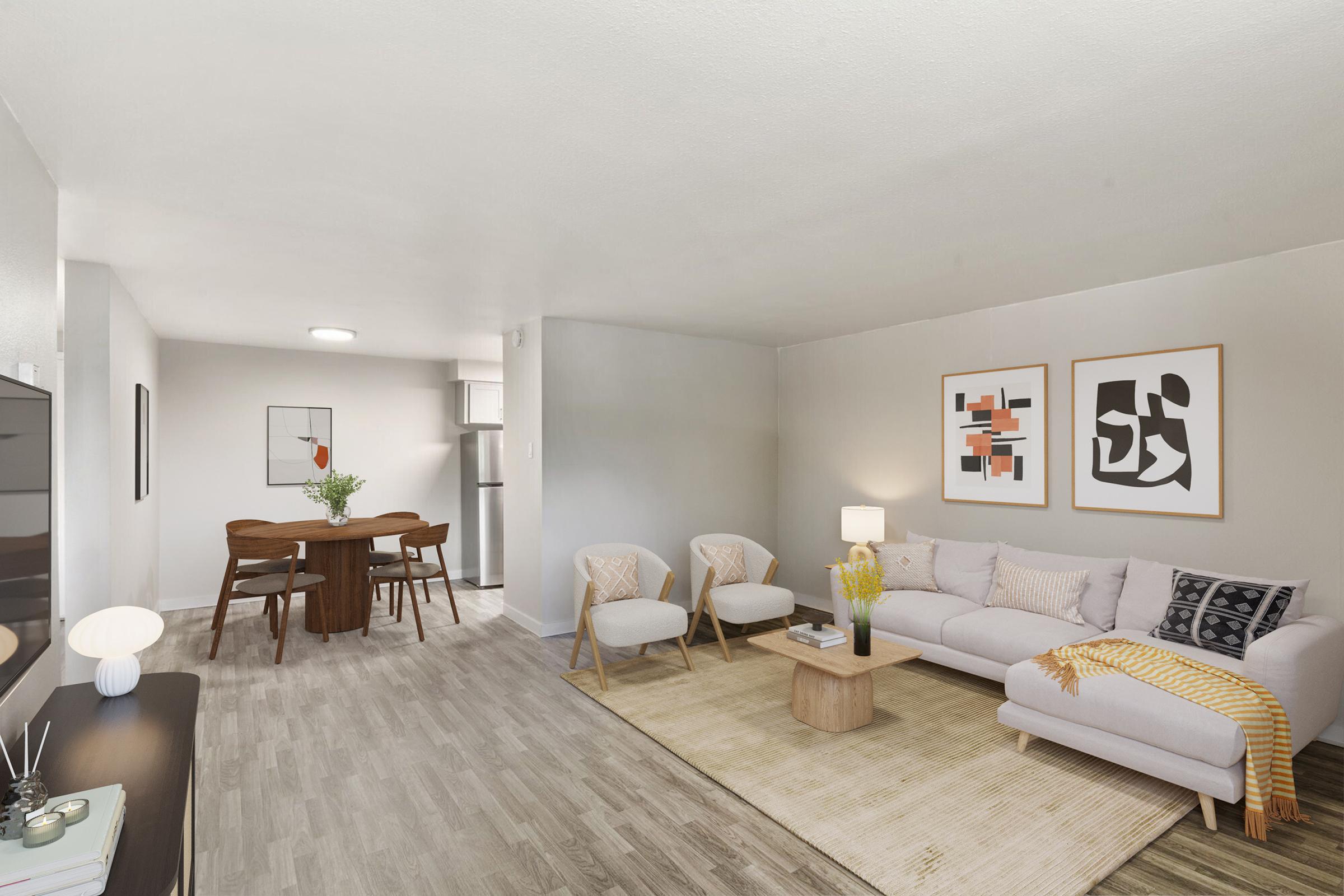 A modern living room featuring a light-colored sofa with decorative pillows, a circular area rug, and two accent chairs. There is a wooden dining table with chairs in the background, along with framed wall art. The space is well-lit and has a neutral color palette, creating a cozy atmosphere.