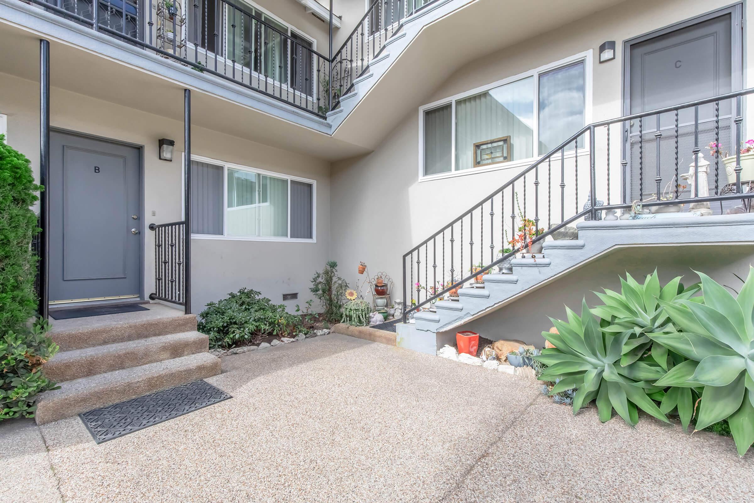 An exterior view of an apartment complex entrance featuring a gray door labeled "B." The area is landscaped with plants and decorative items, a staircase leading to the upper level, and a small garden bed along the wall.