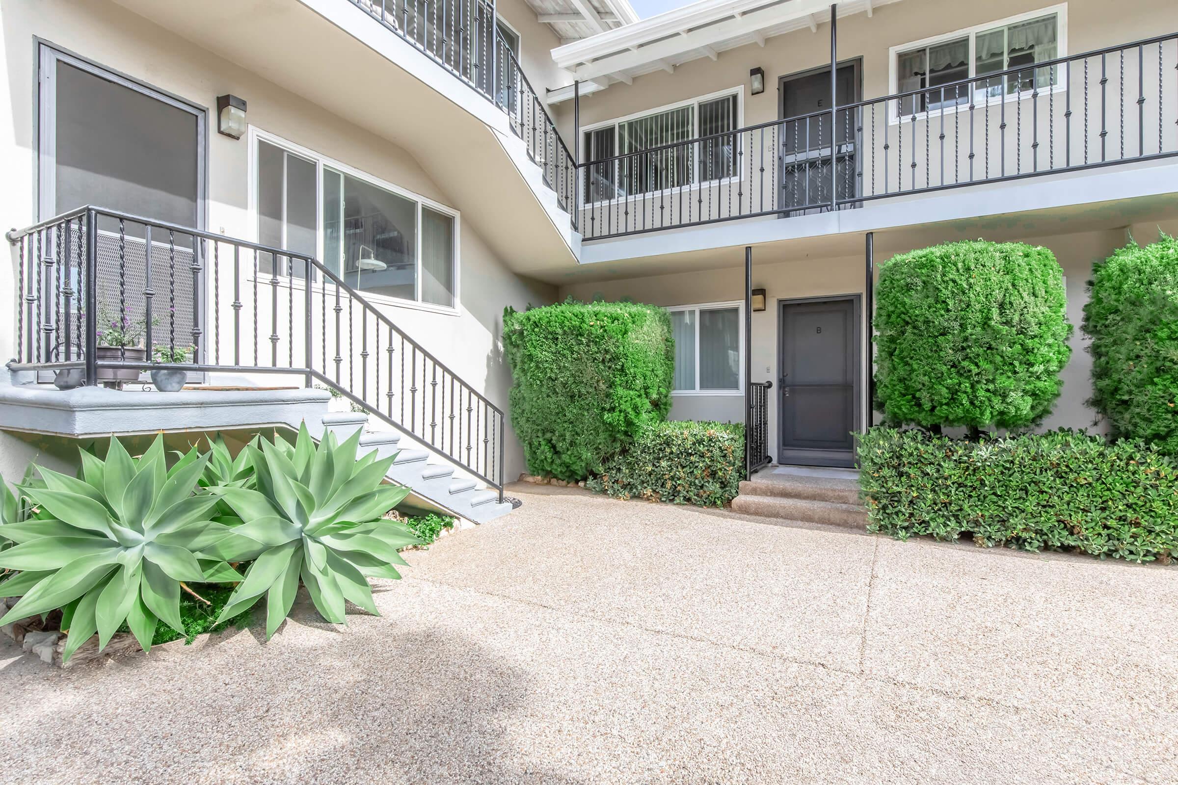 A modern apartment entrance featuring a staircase with black railings, flanked by neatly trimmed hedges. The pathway is made of pebbles, leading to a black front door. Large green succulents add a touch of greenery, enhancing the inviting atmosphere of the entrance area.