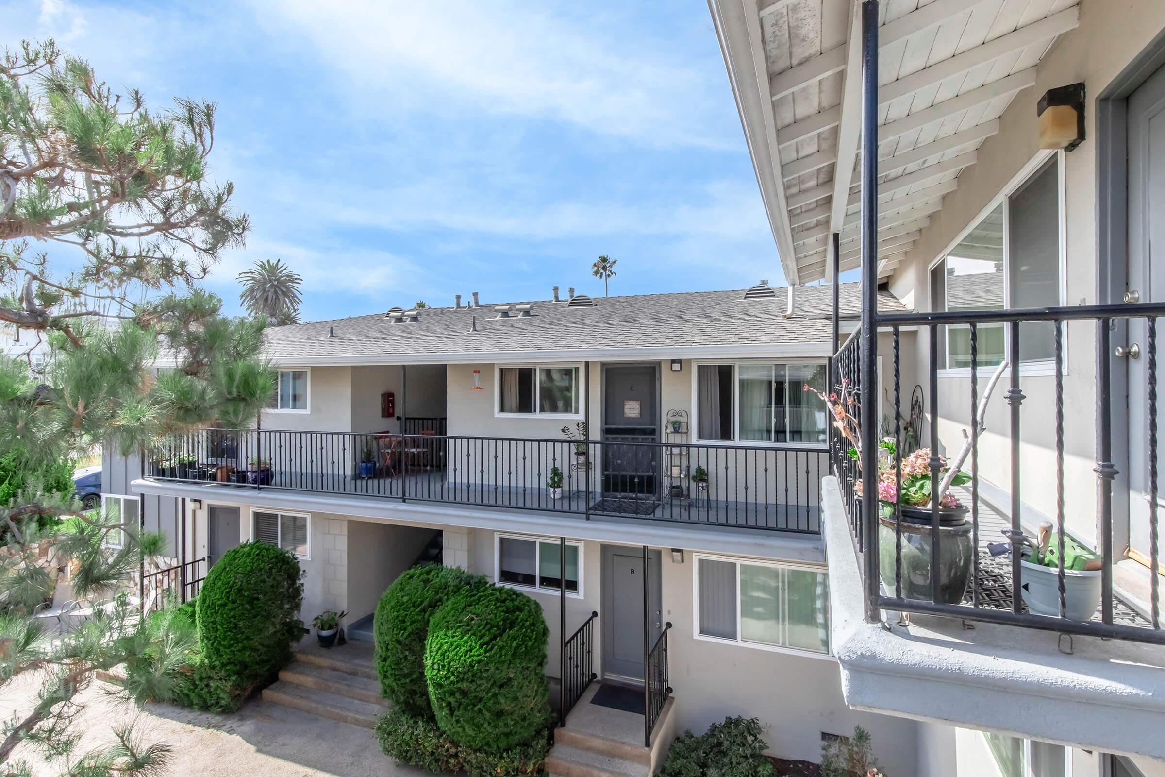A view of a two-story apartment building with balconies, surrounded by greenery. The building features multiple units, each with a front door and small outdoor area. In the background, there are palm trees and a clear blue sky.