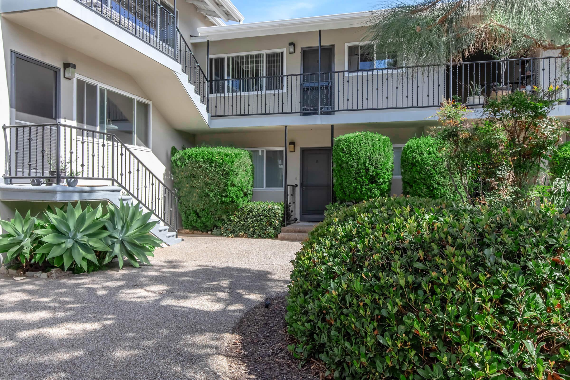 A view of a landscaped courtyard with a path leading to an entrance, flanked by green shrubs and plants. Surrounding two stories of balconies are visible, offering access to adjacent rooms in a residential building. Sunlight casts shadows on the pathway, enhancing the tranquil atmosphere.