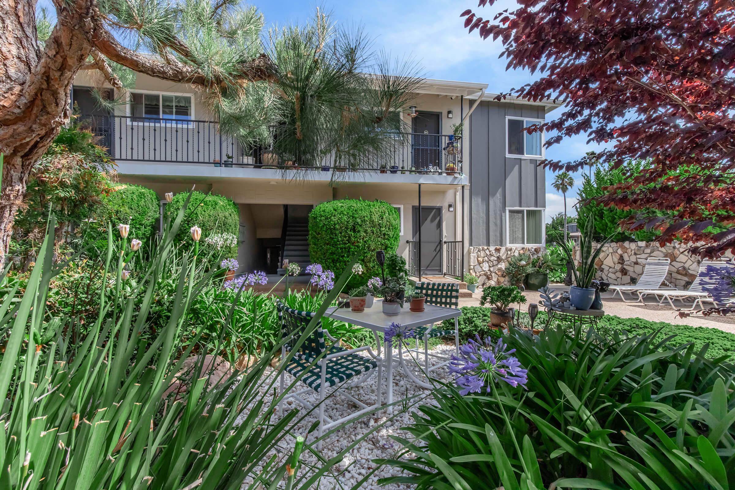 A serene outdoor area featuring a small white table and chairs surrounded by lush green plants and purple flowers. In the background, a two-story building with balconies is partially shaded by trees. Soft sunlight casts a warm glow over the scene, creating a peaceful atmosphere.