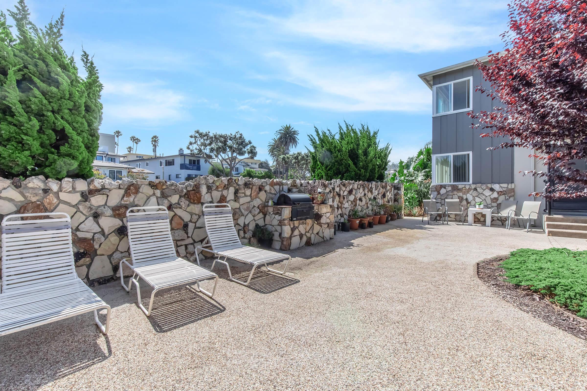 A sunlit outdoor space featuring several lounge chairs arranged near a stone wall. There are potted plants, a grill, and a well-maintained garden area beside a modern building. The scene is bright, with clear blue skies and palm trees visible in the background.