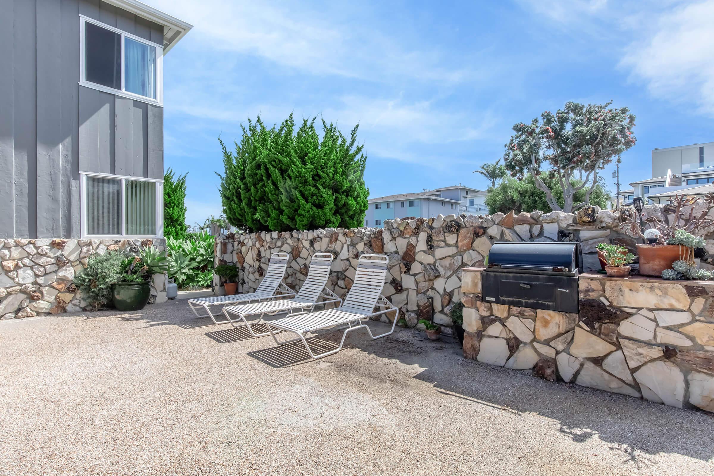 A sunlit outdoor area featuring three lounge chairs arranged on gravel, with a stone wall in the background. A barbecue grill is positioned nearby, surrounded by potted plants and shrubs, set against a clear blue sky. The scene conveys a relaxing, inviting atmosphere.