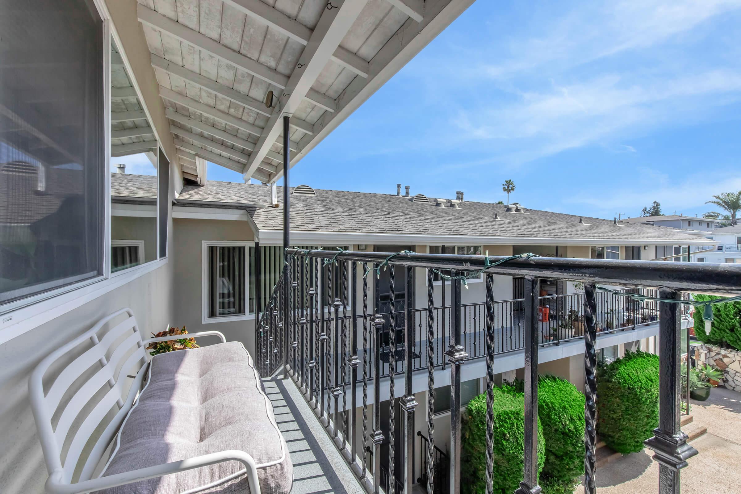 A balcony view showcasing a white bench with cushions, overlooking a courtyard with green hedges. The scene features modern apartment buildings under a clear blue sky, with a charming architectural design and a hint of palm trees in the background.
