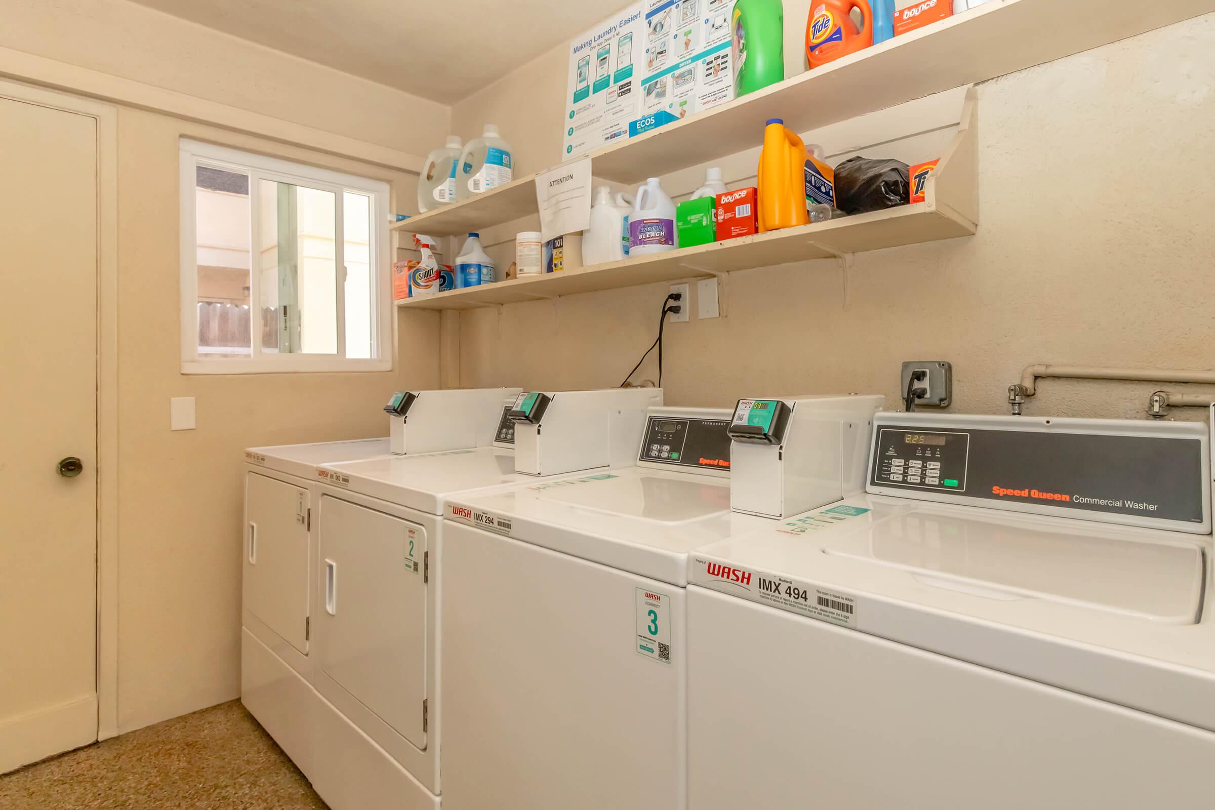 A clean laundry room featuring three commercial washing machines and a shelf stocked with various laundry supplies, including detergents and fabric softeners. There is a window allowing natural light and a door leading to another area. The overall setting is tidy and organized.