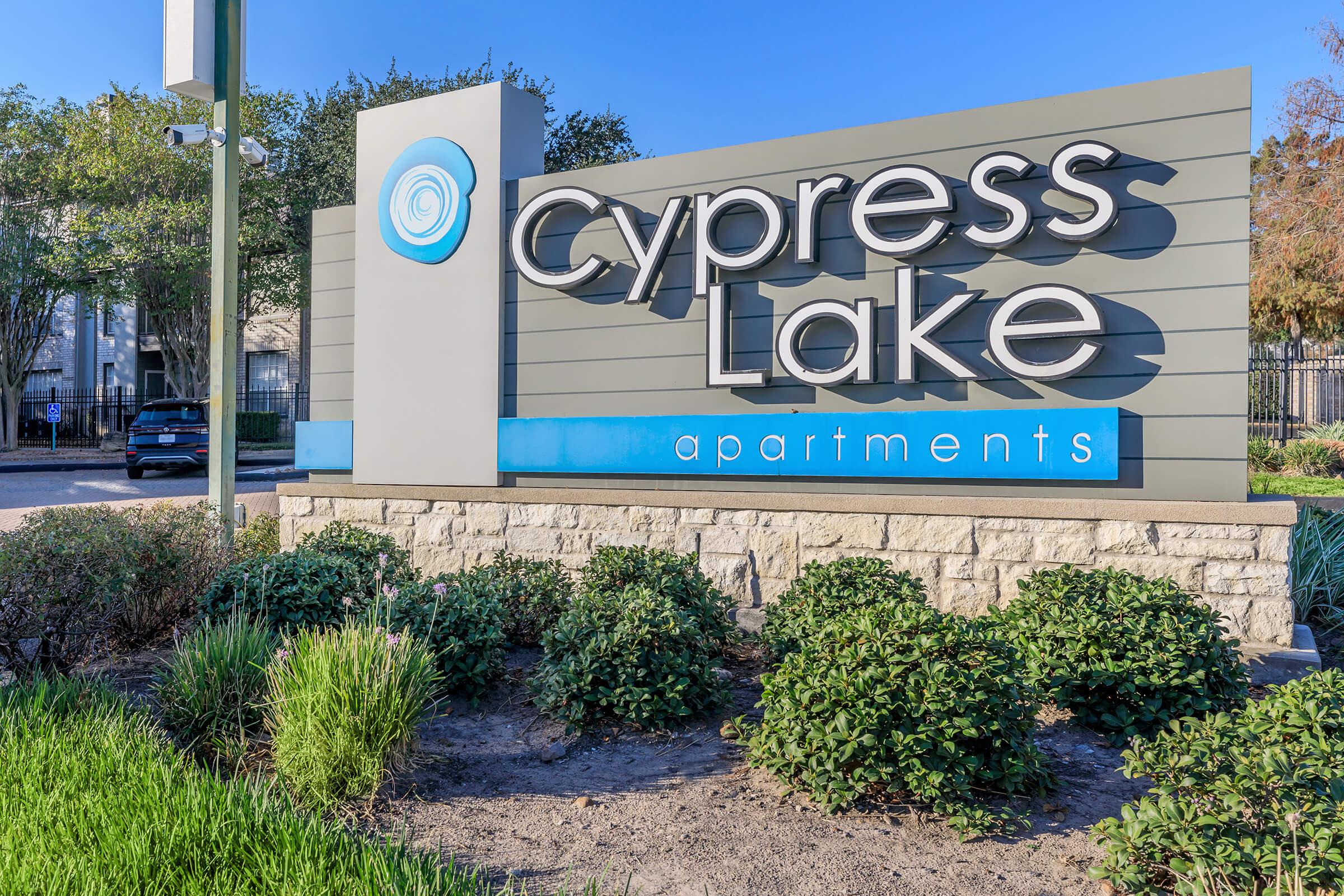 Sign for Cypress Lake Apartments featuring a modern design with the name "Cypress Lake" prominently displayed. The sign includes a circular logo and is surrounded by landscaping, including green shrubs and a clean pathway. The background shows a glimpse of the apartment buildings.